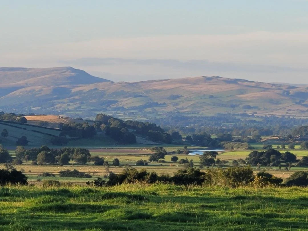 Scenic view of the rolling hills of Lancaster and the Lake District mountains in the distance with a lush green field in the foreground, partly cloudy sky.
