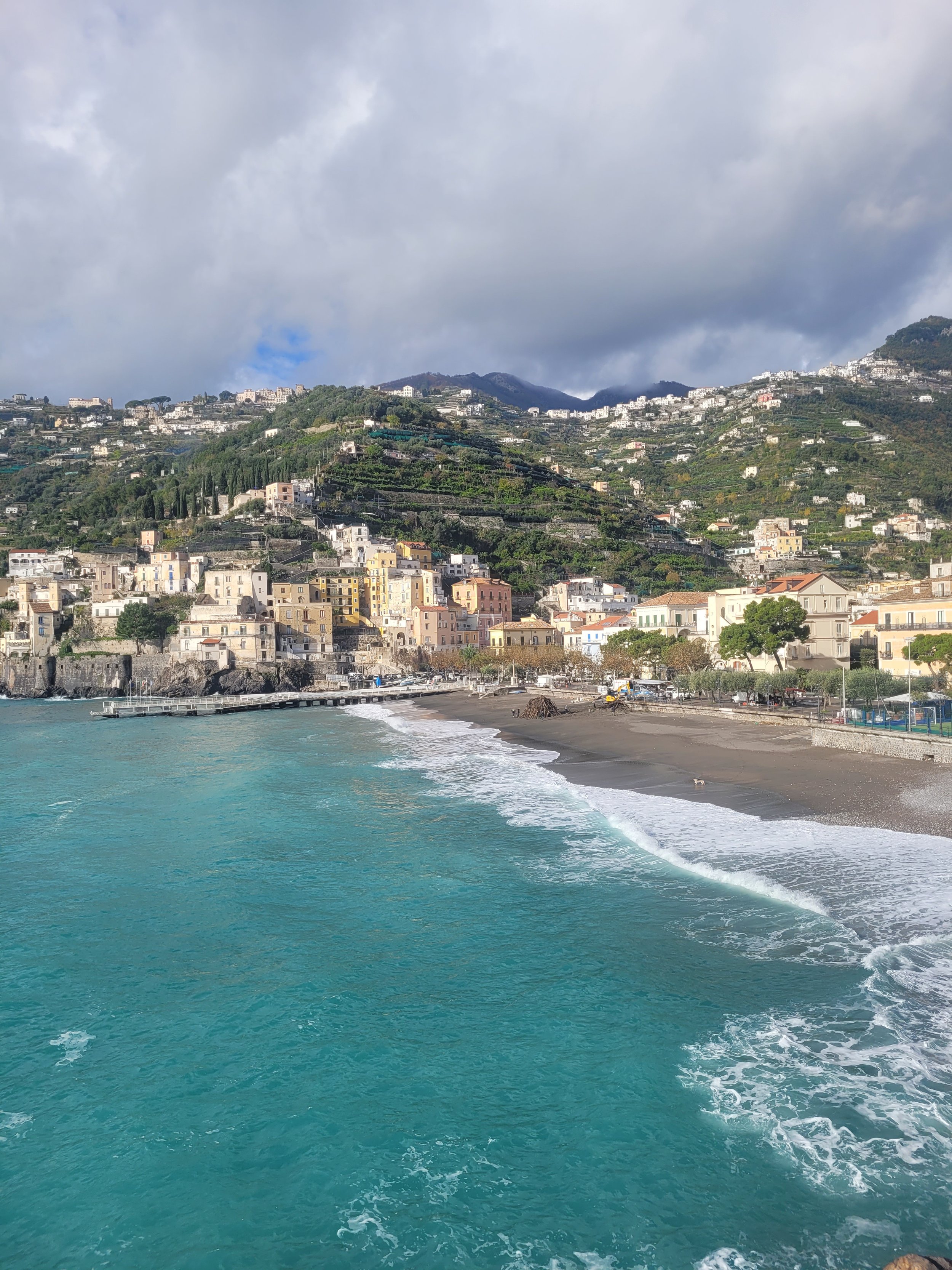 A coastal scene showing a beach with dark sand, turquoise waters, and waves. Behind the beach, there are colorful buildings along the shoreline, and a hillside with houses and greenery rising in the background under cloudy skies.