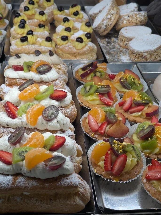 Assorted éclairs topped with fruit and cream, and fruit tarts with colorful sliced fruits on a pastry base.
