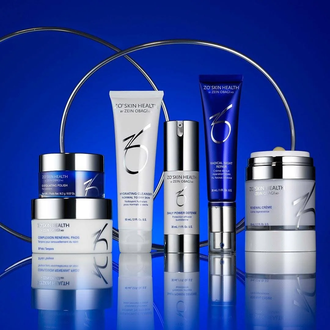 Collection of skincare products including cleansing, renewal pads, defense, night repair, and crème from ZO Skin Health by Zein Obagi, arranged with metal hoops on a reflective blue surface.