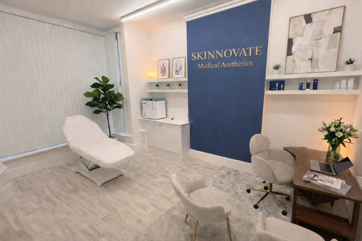 Interior of a medical aesthetics clinic reception area with a treatment bed, a large potted plant, a wooden desk with a laptop and flower vase, and decorative shelving on the wall.