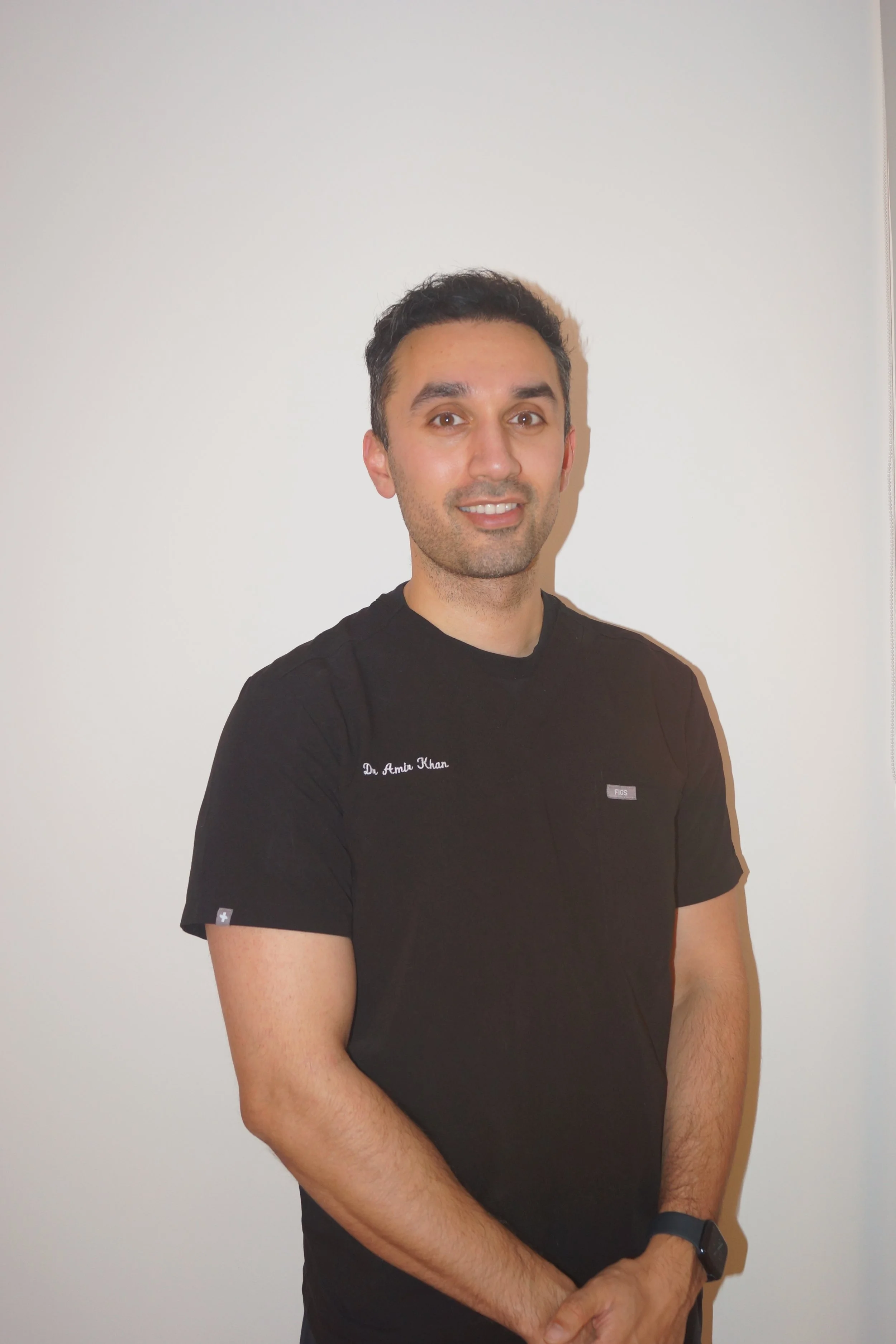 A man standing against a plain white wall, wearing a black t-shirt with the name tag 'Dr. Amir Khan' and a black wristwatch. He has short dark hair, a light complexion, and is smiling at the camera.