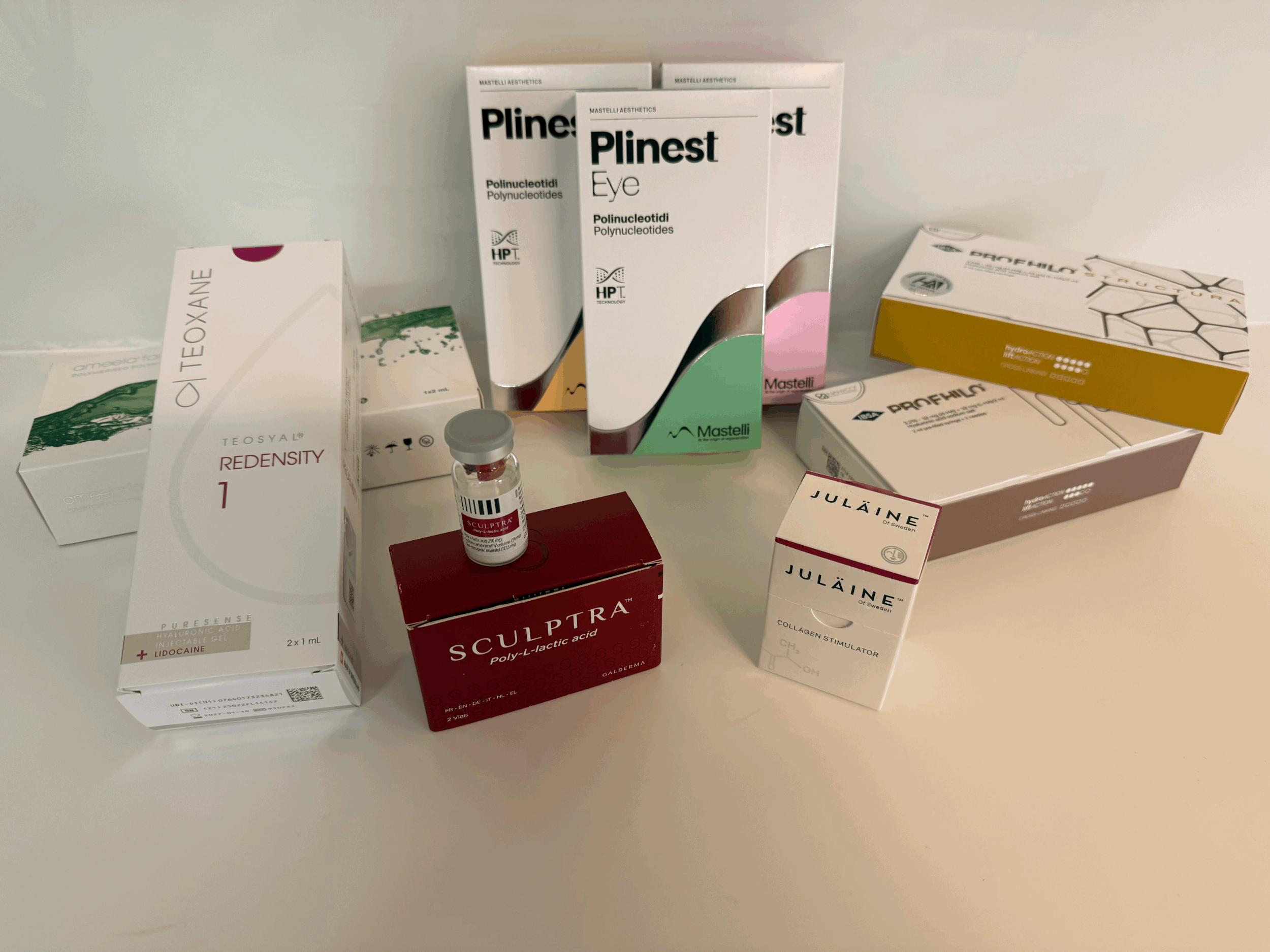 Various cosmetic and medical products on a white table, including boxes labeled Teoxane, Plinest, Sculptra, Julaine, and other containers, some with syringes and vials.