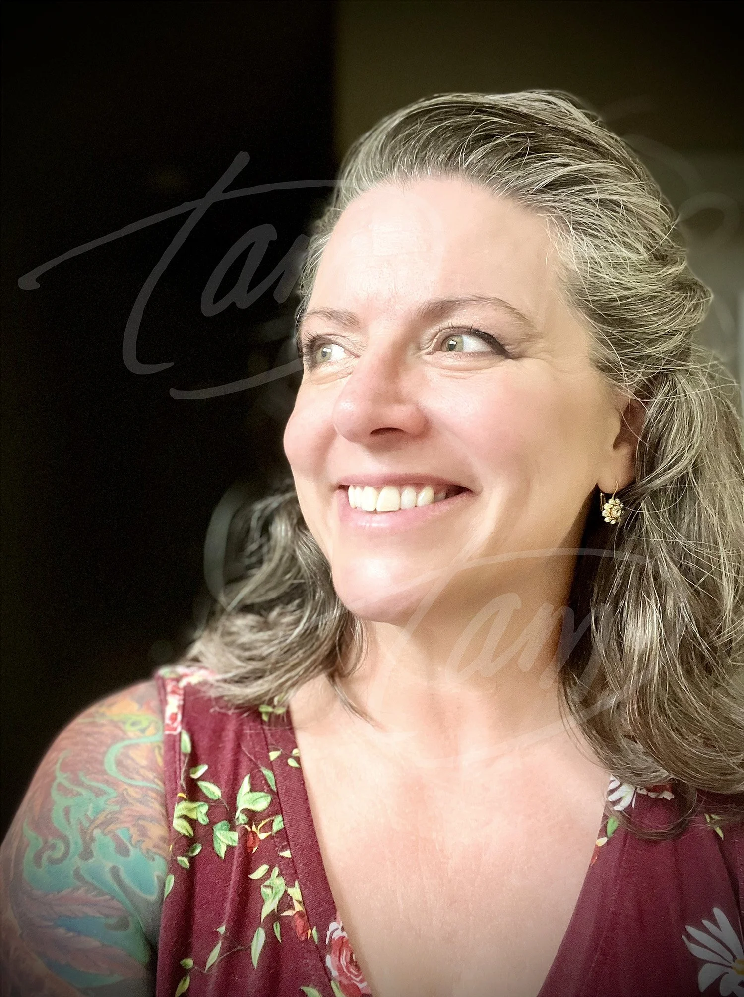 Artist Tam, smiling, wearing a maroon floral sleeveless top, gold earrings, and a colorful tattoo showing on her left shoulder.