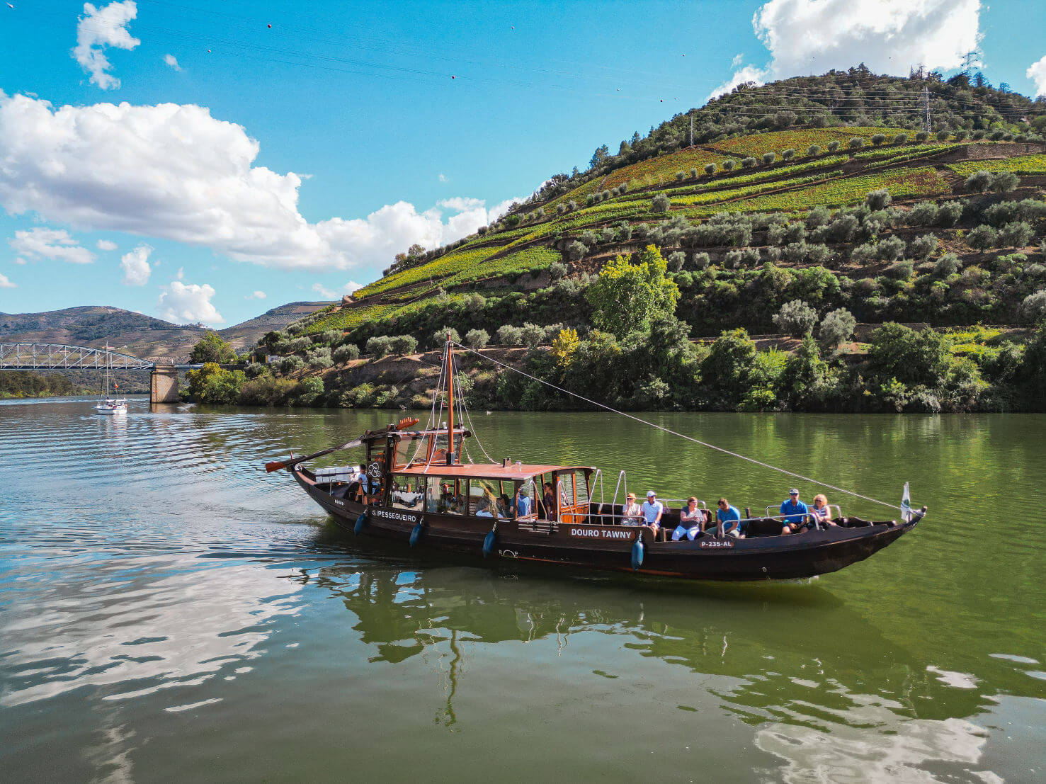 Traditional rabelo boat carrying passengers on the Douro River alongside terraced hillsides.