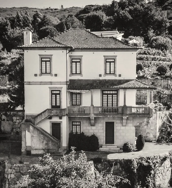 A three-story house with a tiled roof, multiple windows, and a balcony on the second floor, situated on a hillside with trees and shrubs in the background.