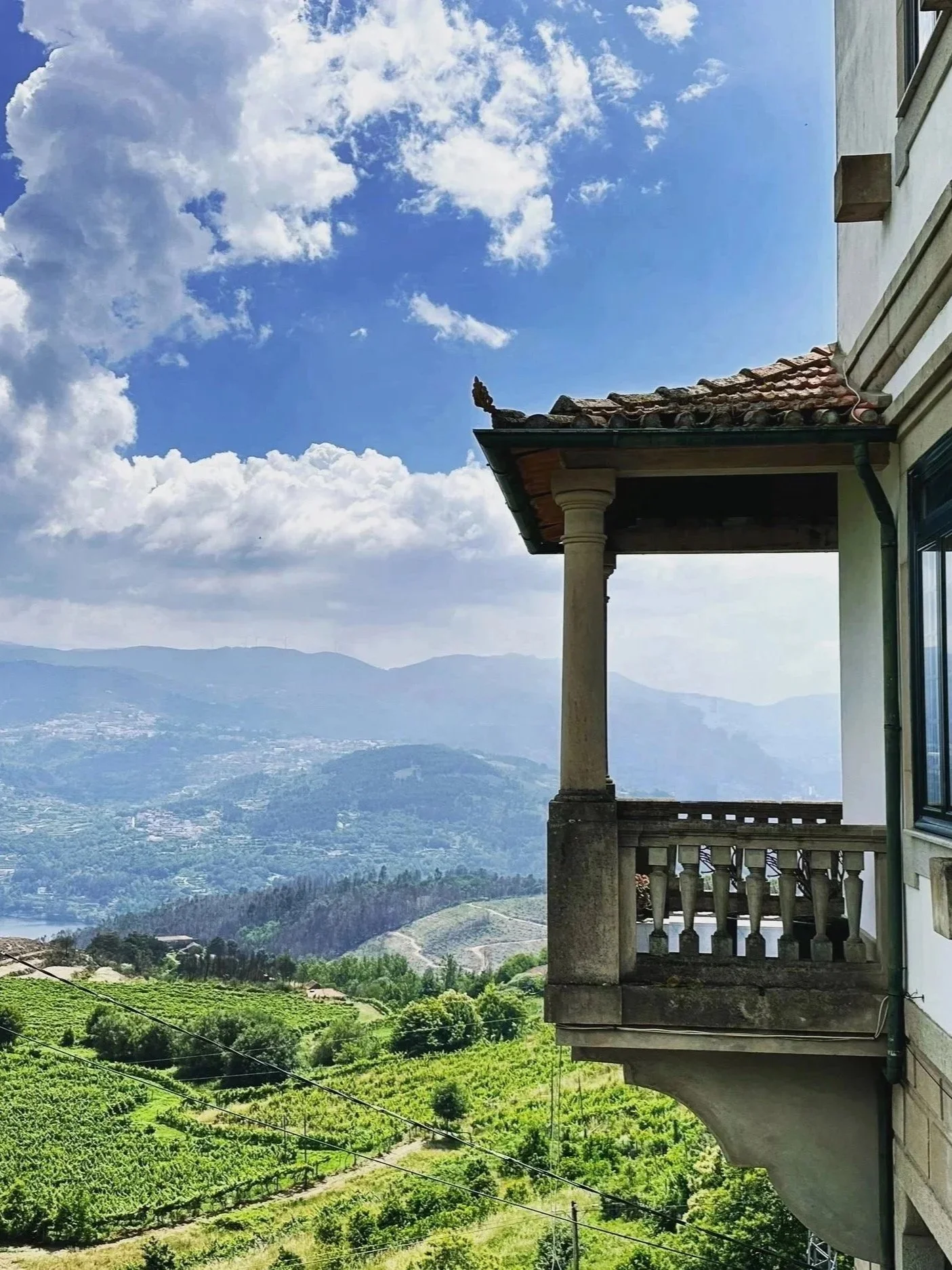View of mountains and a vineyard from a balcony with stone columns and a tiled roof.