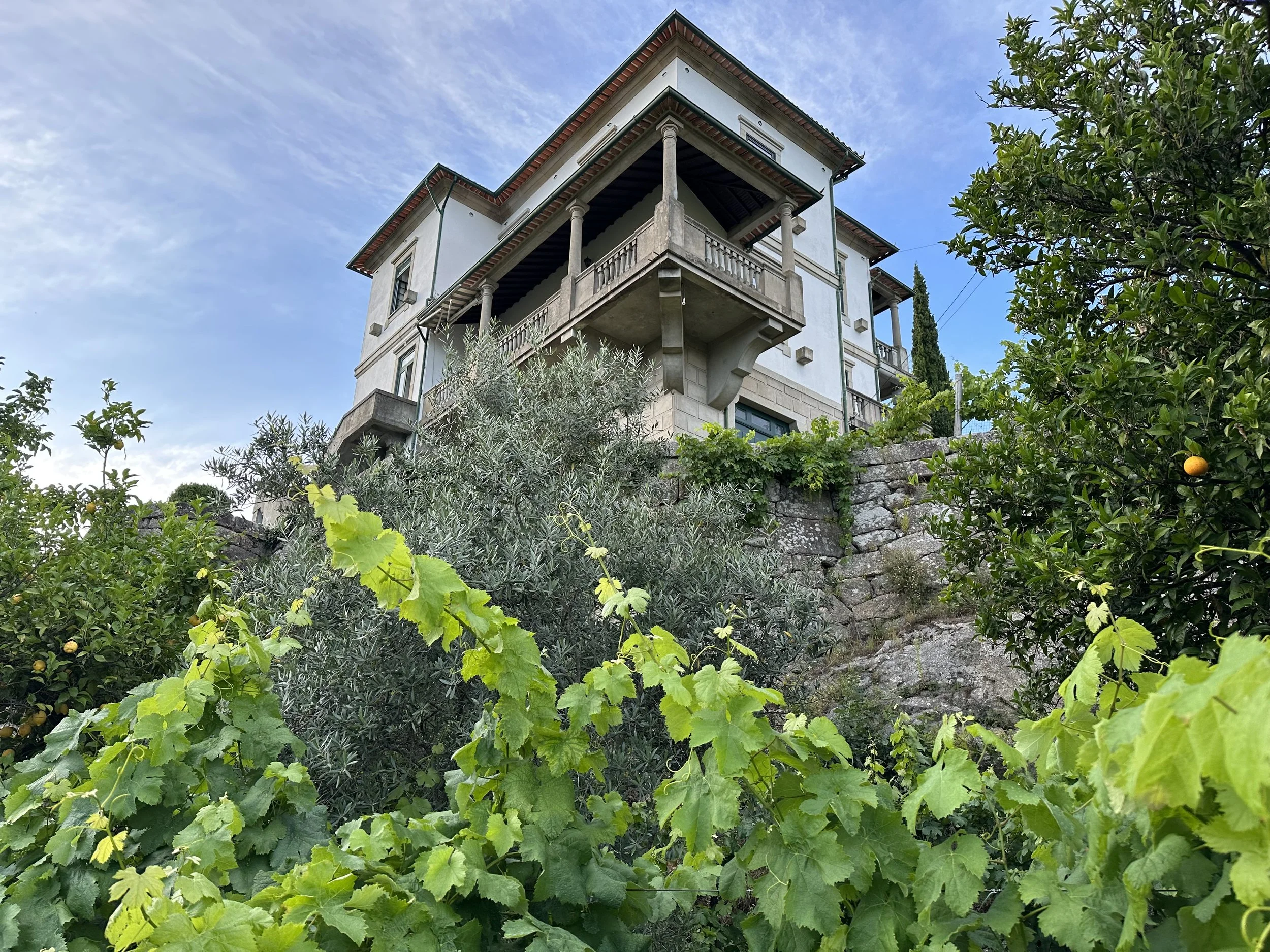 A multi-story house on a hillside surrounded by greenery, with a stone retaining wall and visible balcony.