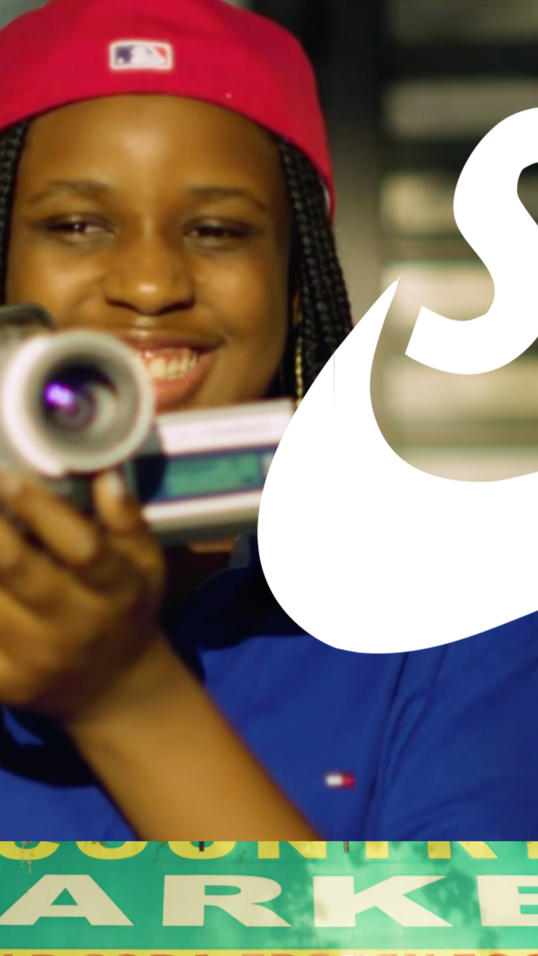 A woman wearing a red cap holding a video camera and smiling at the camera.