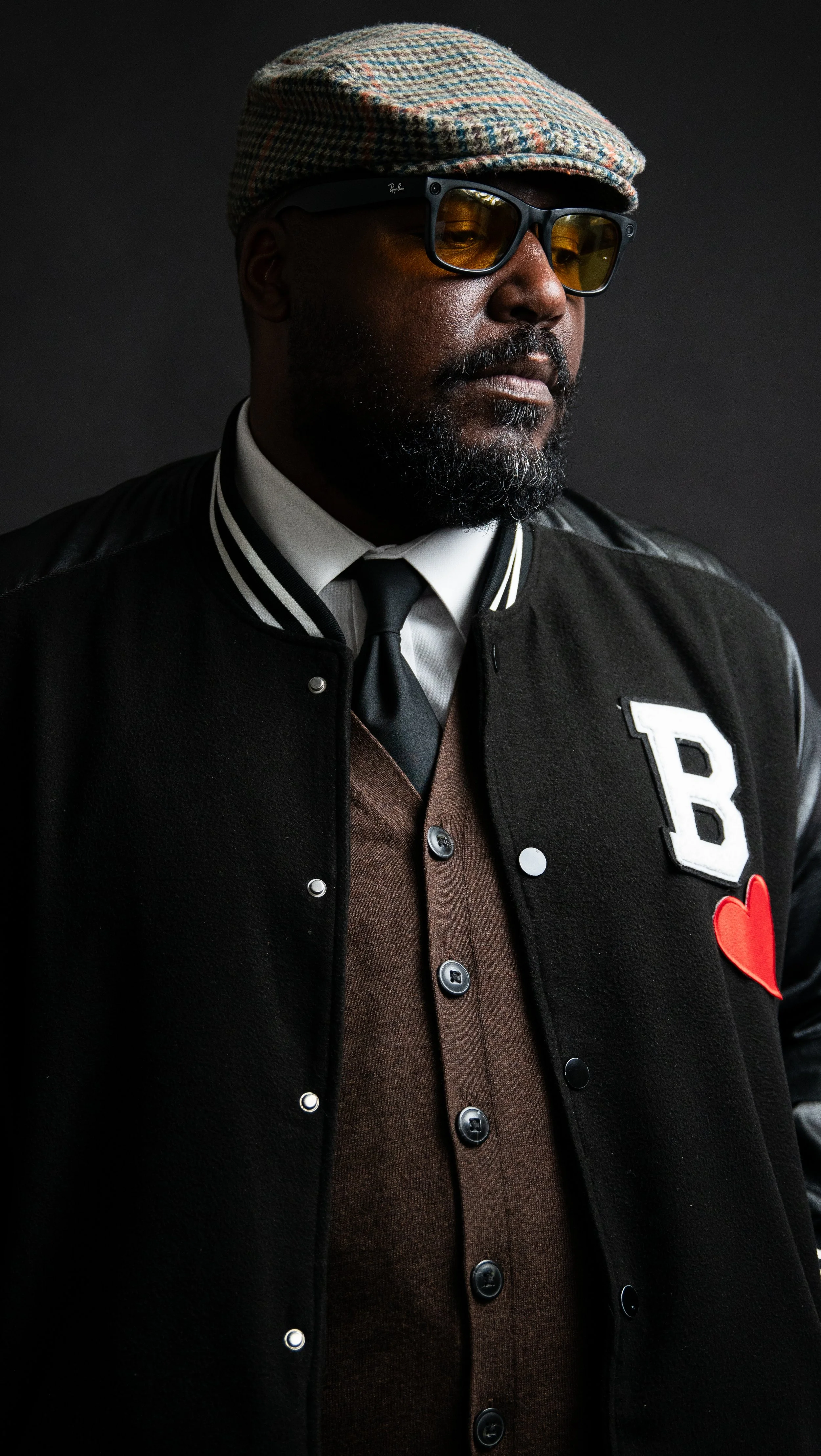 A stylish man in a brown sweater, black jacket with a letter patch and red heart, white shirt, and black tie, wearing sunglasses and a patterned flat cap, standing against a dark background.