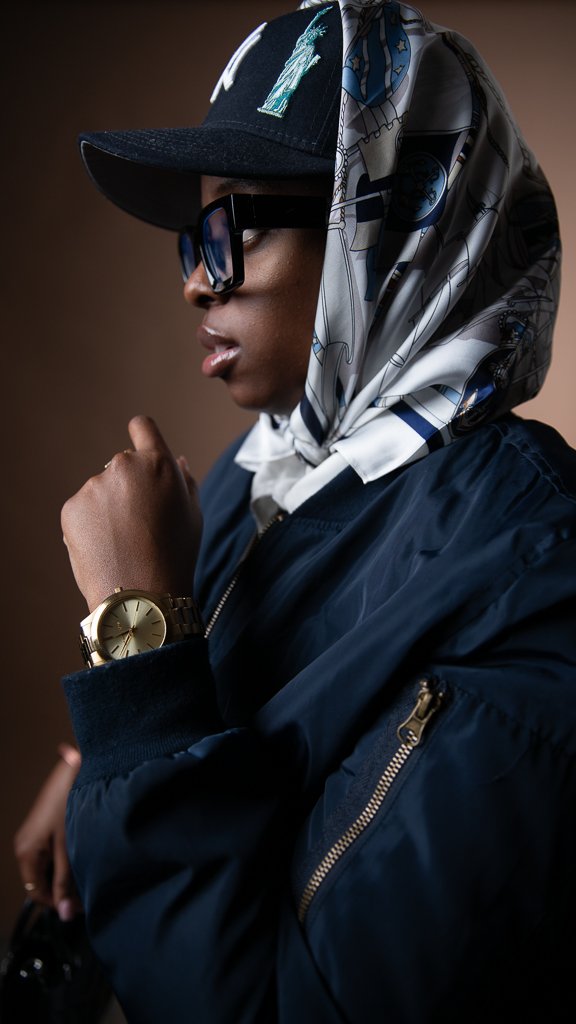 A woman wearing a black cap, glasses, a silk scarf, a dark blue bomber jacket, and a gold wristwatch.