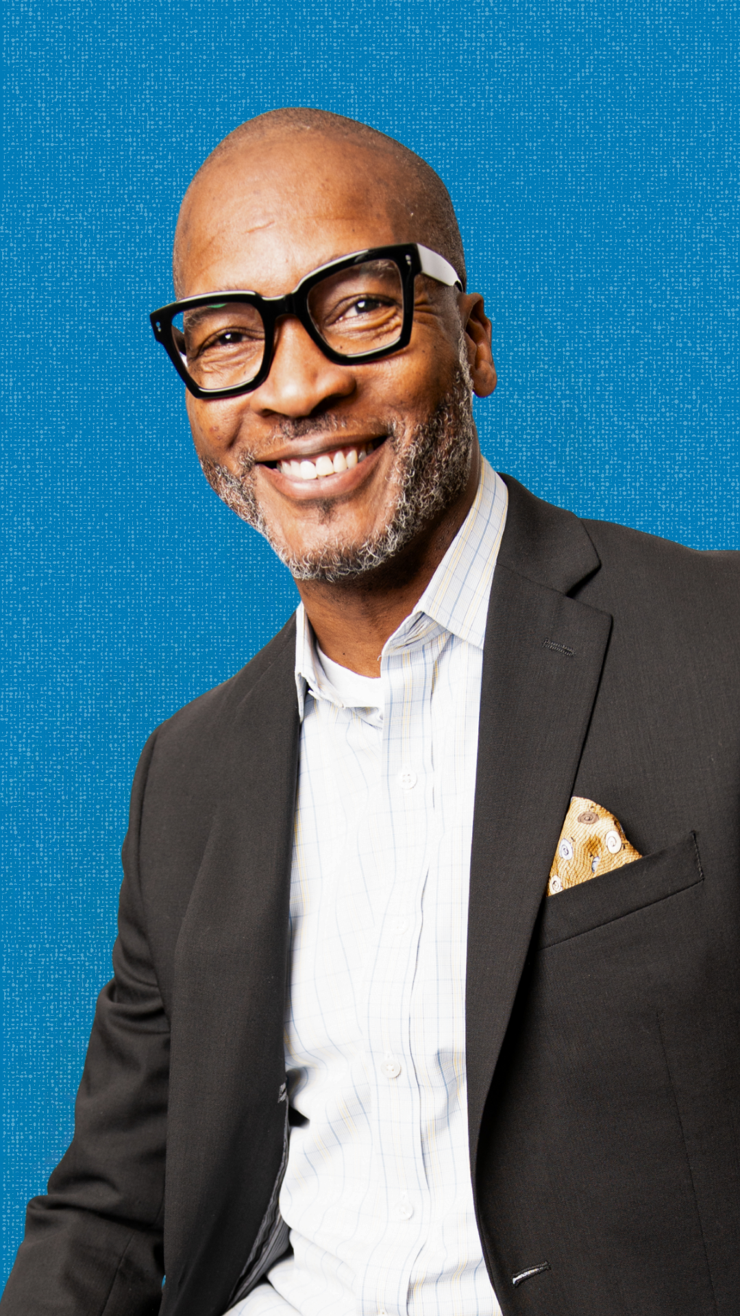 A smiling middle-aged man with a gray beard, wearing black glasses, a black suit, a white checkered shirt, and a pocket square, posing against a blue textured background.