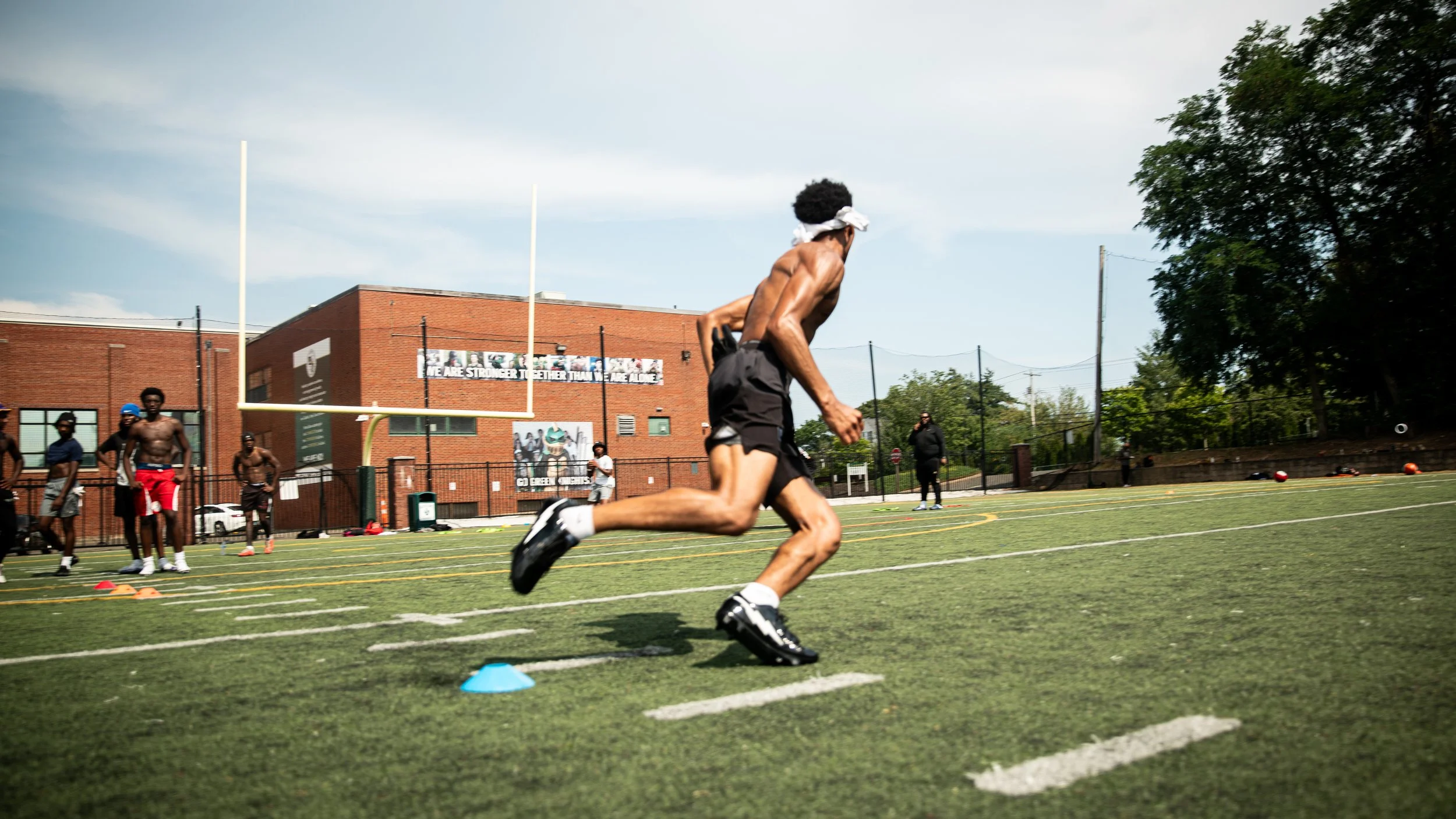 A shirtless man with a cloth tied around his head running on a football field during a training drill, with other shirtless men and a coach in the background.