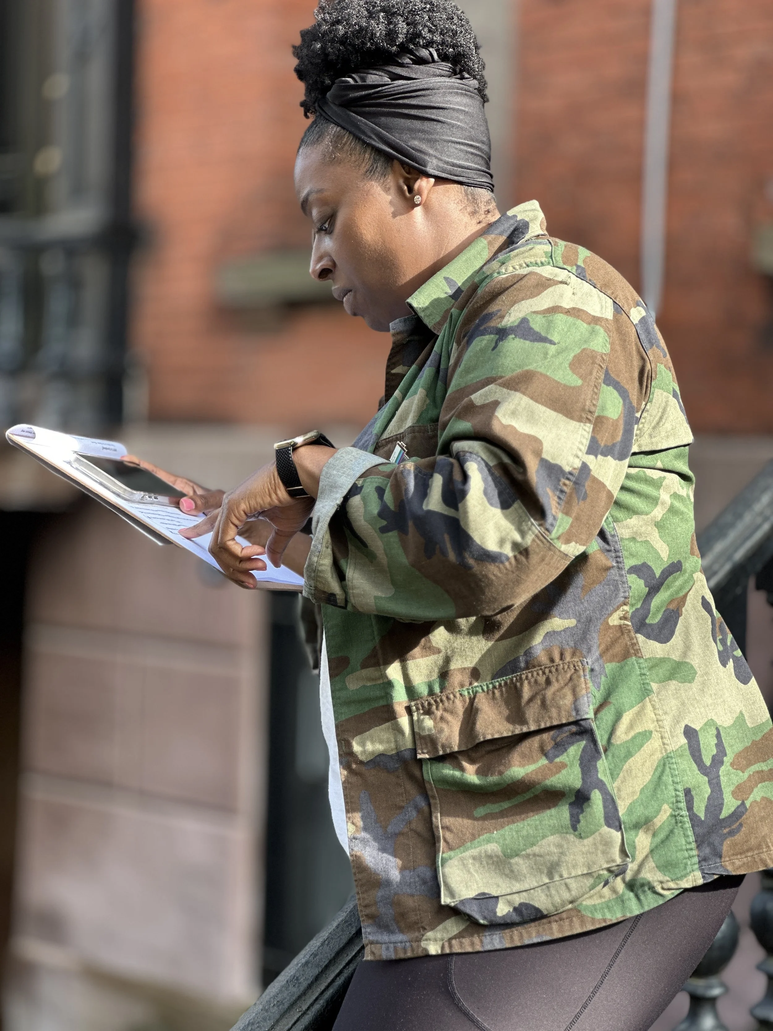 A woman in a camouflage jacket looking at her phone outdoors in front of a red brick building.