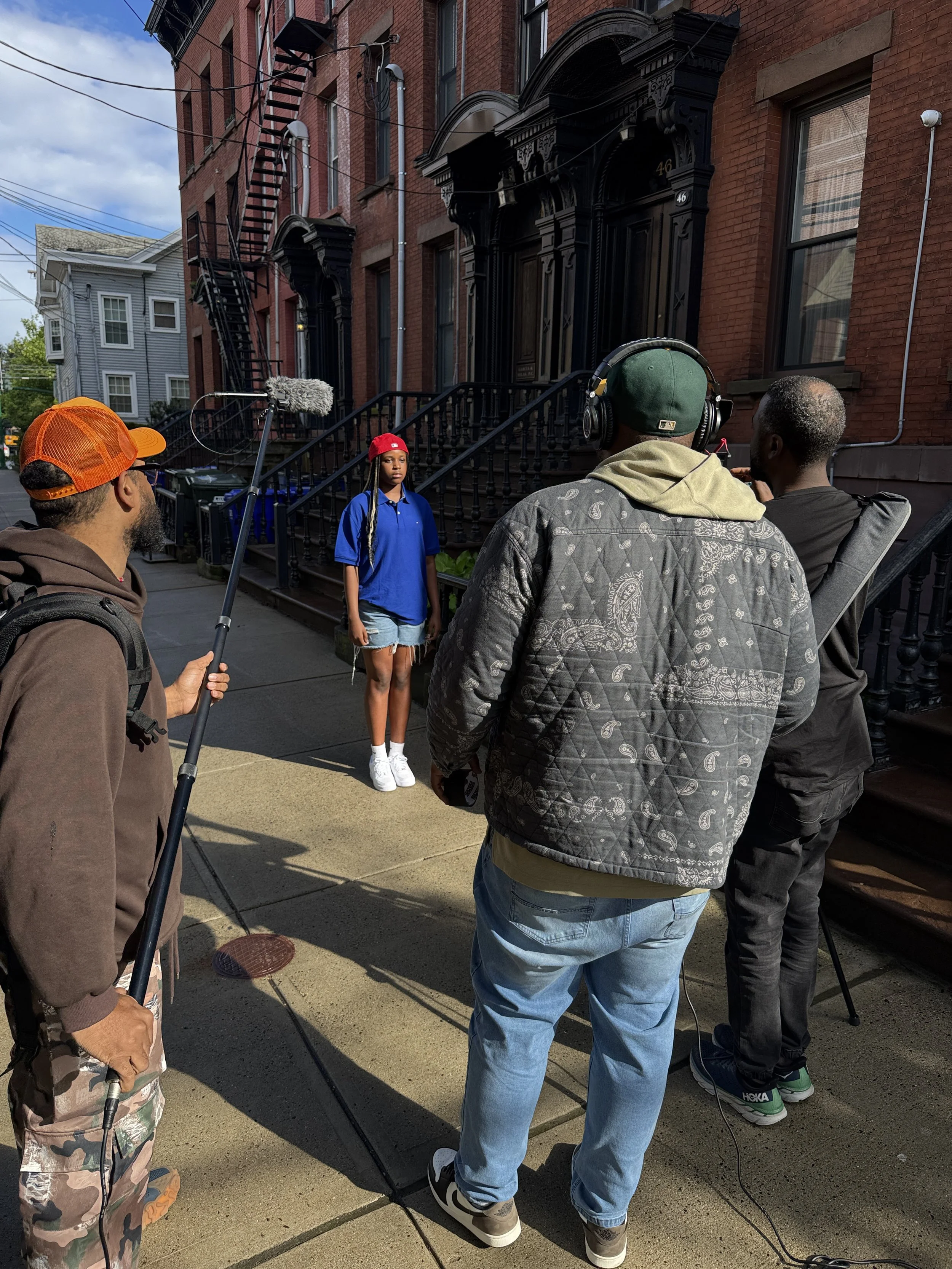 A film crew filming a young woman standing on the sidewalk in front of a brick apartment building, with crew members recording and directing her.