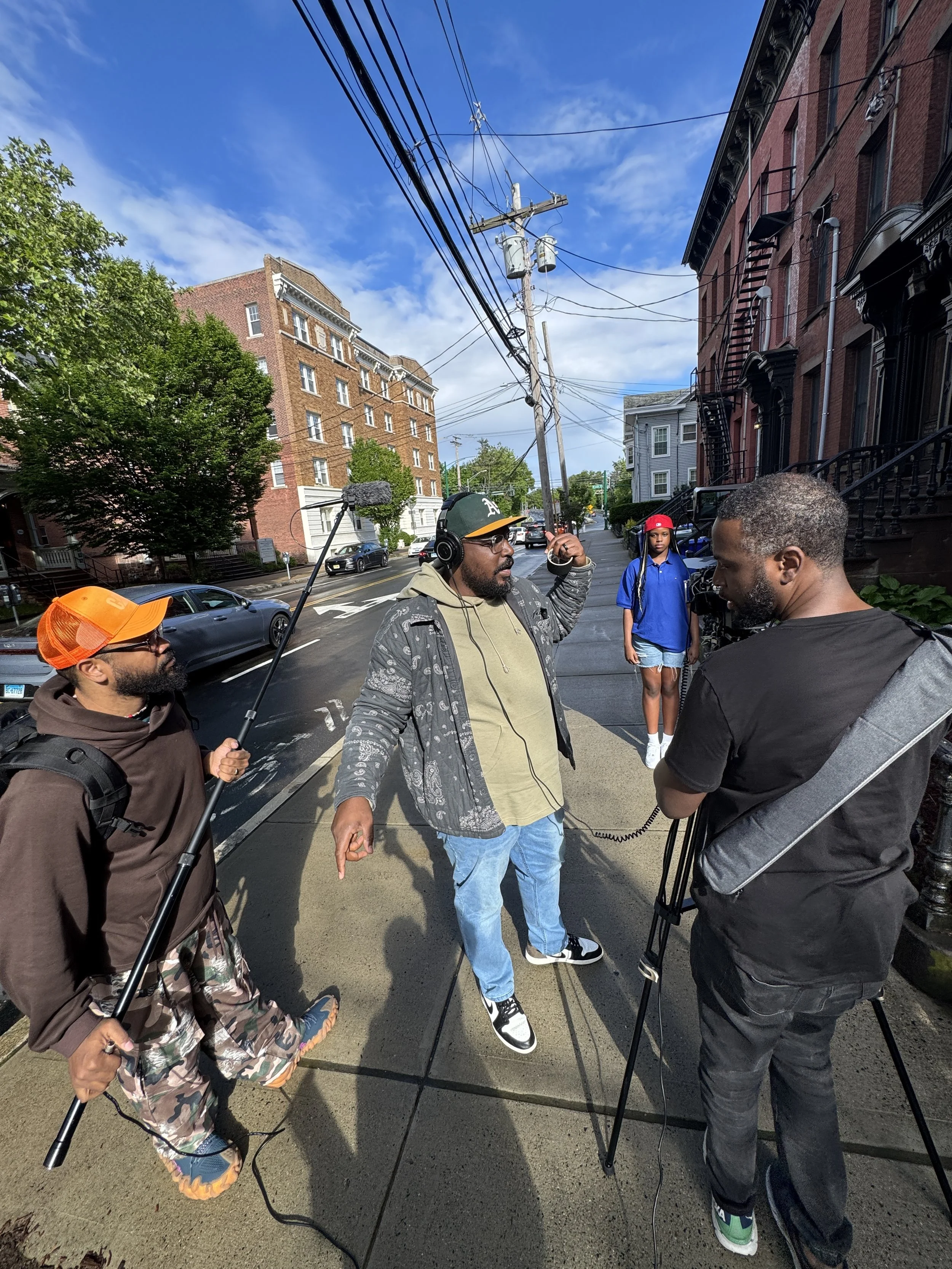 Group of people filming a street scene with a man speaking into a microphone, there is a woman in the background, and the street has cars, buildings, and power lines.