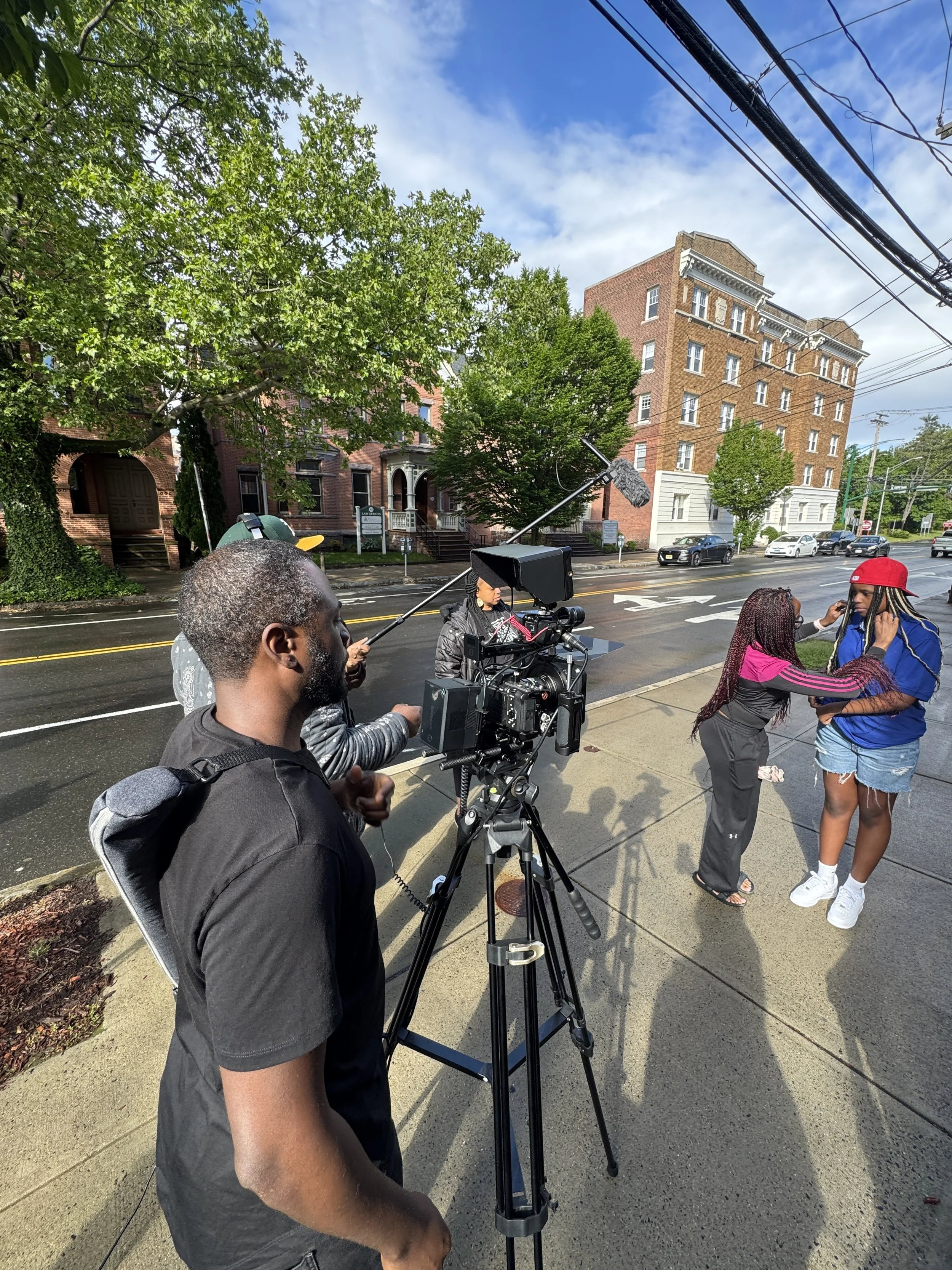 Filming of a scene on a city sidewalk with a camera crew filming two women, one applying makeup or adjusting something on the other, with a professional camera mounted on a tripod, and a boom microphone operator capturing audio, trees and buildings in the background.