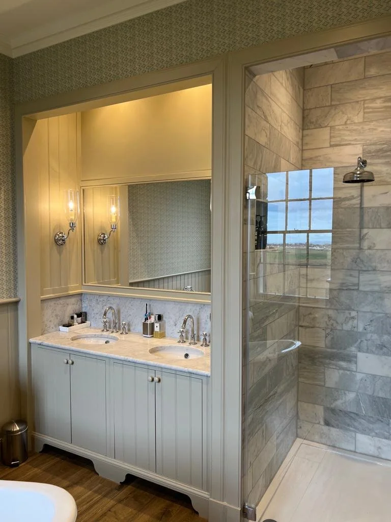 Dual sink bathroom vanity with marble countertop, large mirror, wall-mounted light fixtures, and glass-enclosed shower with marble tile walls.