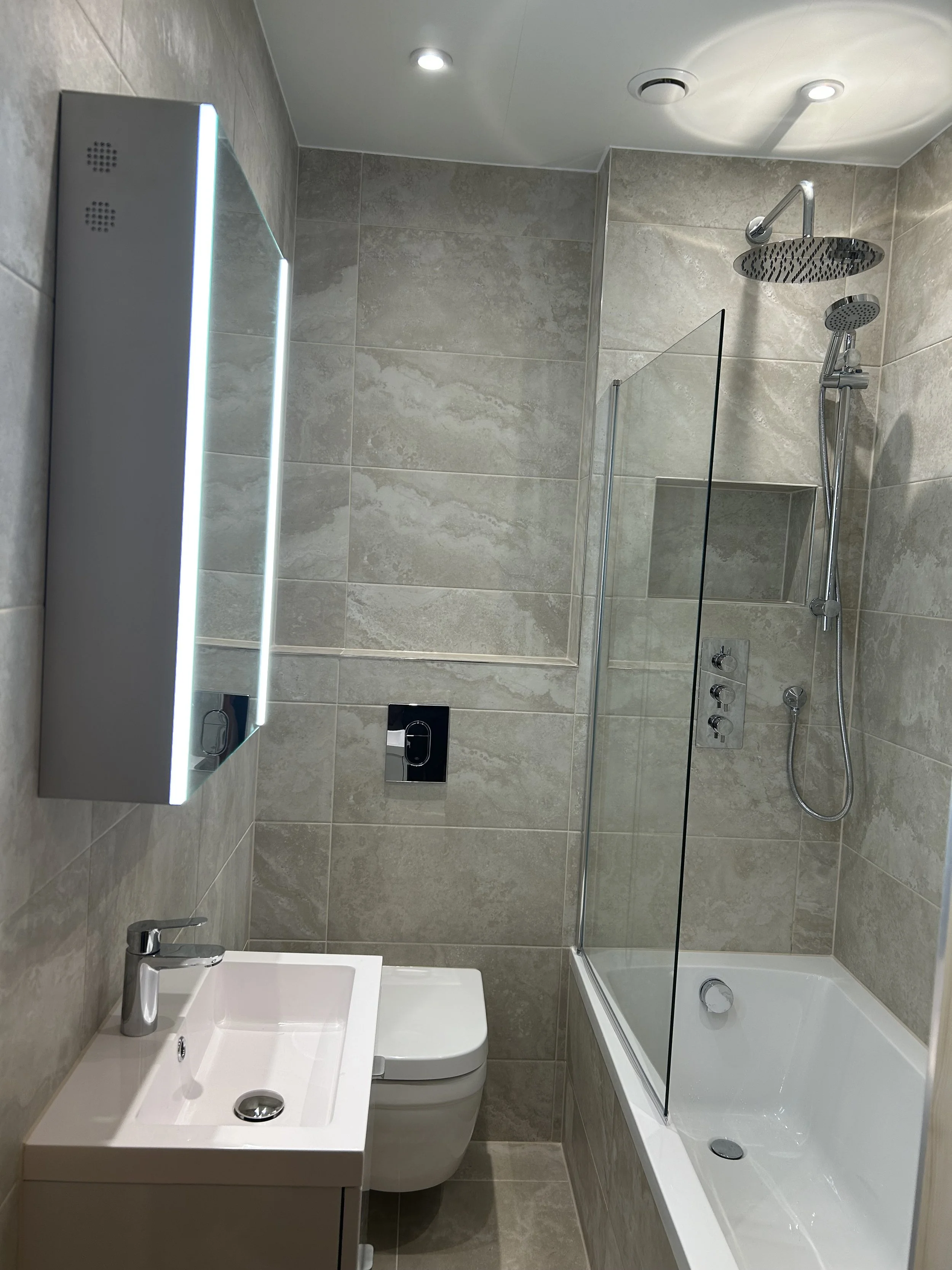 Modern bathroom with beige tiled walls and floor, a white sink with a chrome faucet, a wall-mounted toilet, a glass shower enclosure with a built-in niche, a rainfall showerhead, a handheld shower, and a mirrored cabinet with LED lighting.