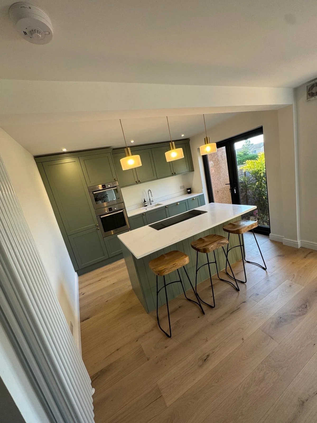 Modern kitchen with green cabinetry, a large island with a white countertop, three wooden bar stools, and hanging yellow pendant lights. There is a sliding glass door leading outside and hardwood flooring.