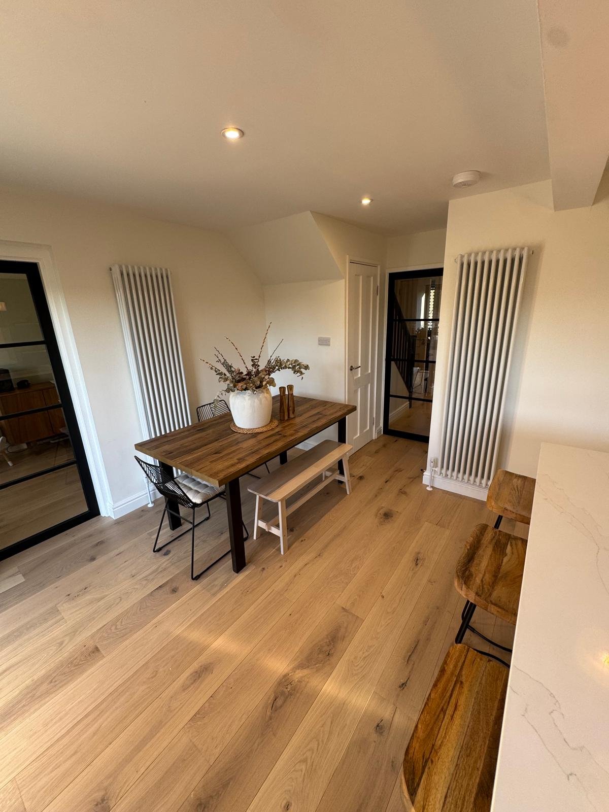 Modern dining room with wooden table, black chair, wooden bench, and bar stools, featuring light wooden flooring and white walls.