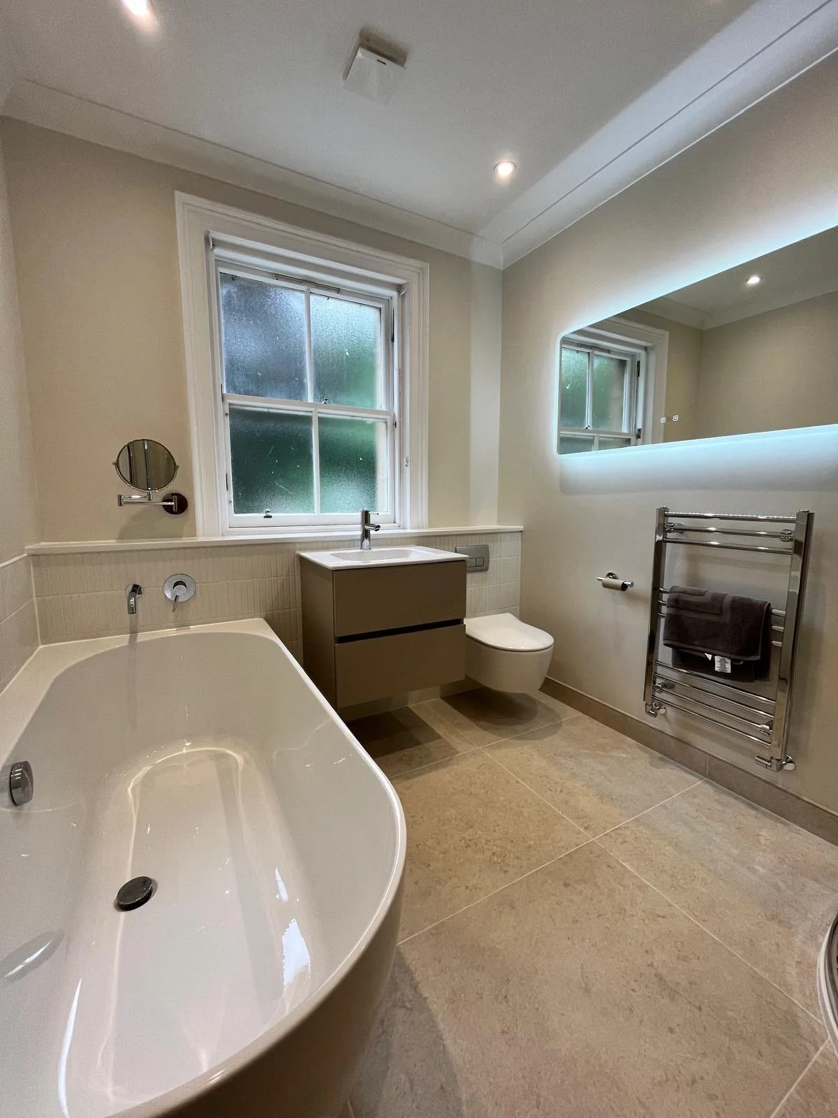 Modern bathroom with a white bathtub, a frosted window, a small vanity with a sink, a mounted mirror, a toilet, and a heated towel rack with a black towel.