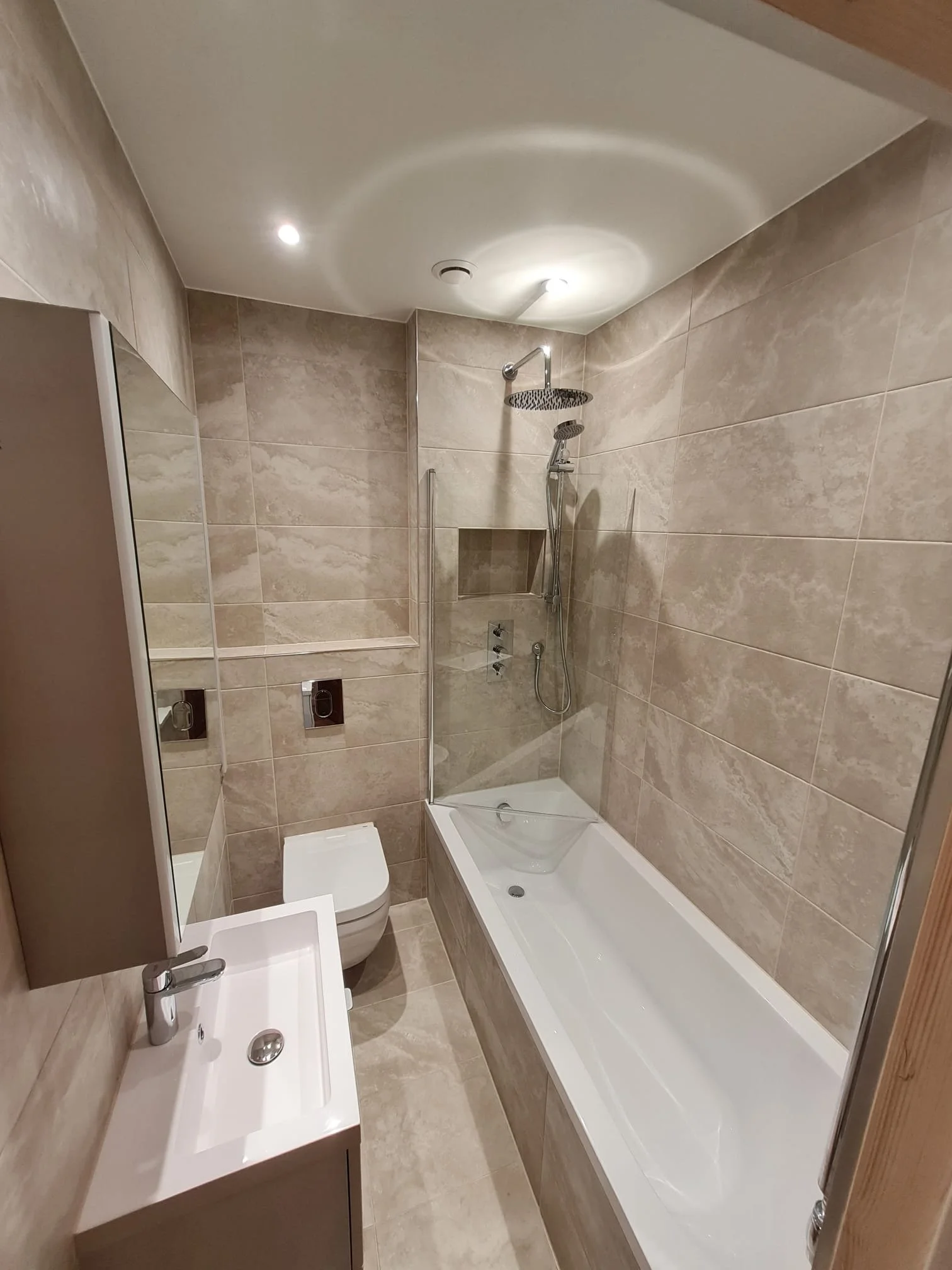Small modern bathroom with beige tiled walls and floor, a white bathtub with a glass shower screen, a wall-mounted toilet, a compact white sink with a chrome faucet, and a mirrored cabinet above the sink.