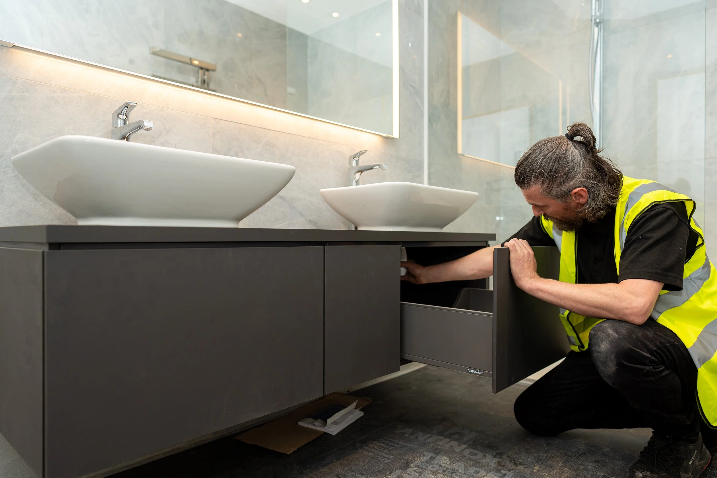 A technician in a yellow safety vest installing or repairing a bathroom vanity drawer beneath two white vessel sinks on a marble countertop, in a modern bathroom with large mirrors.