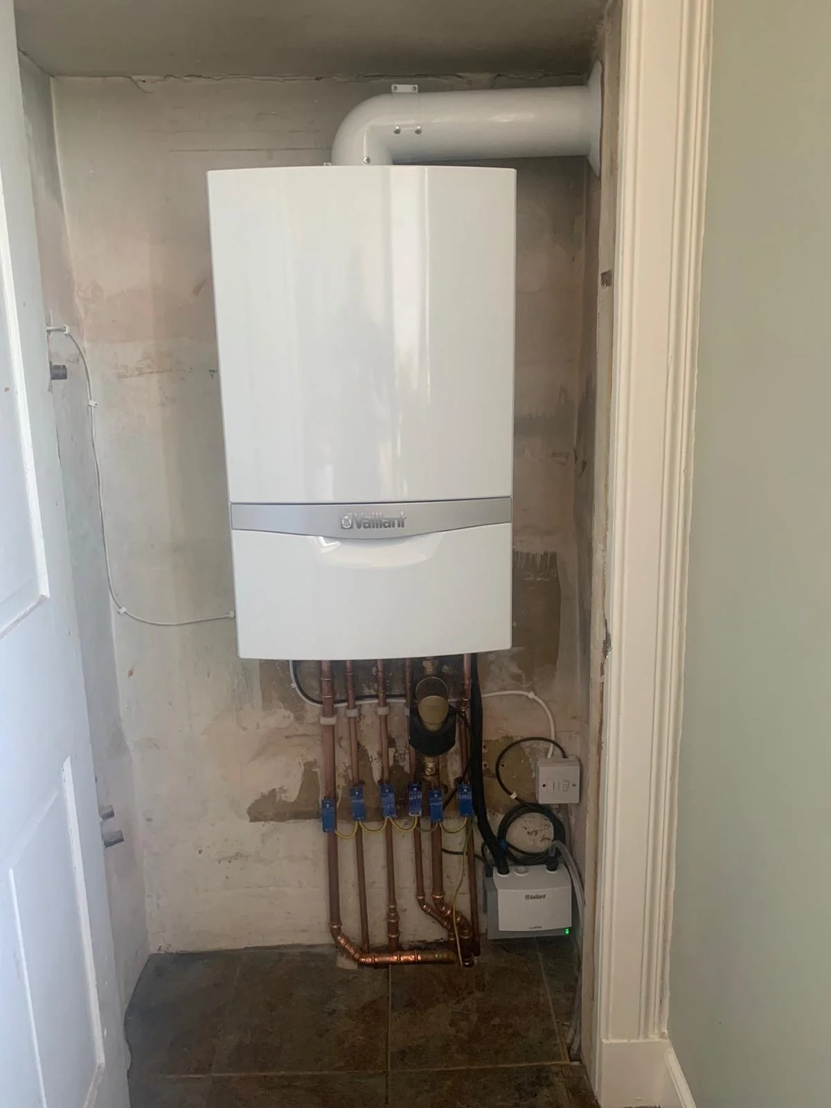 A new white boiler installed on an unfinished wall with exposed pipes and a small control box below.