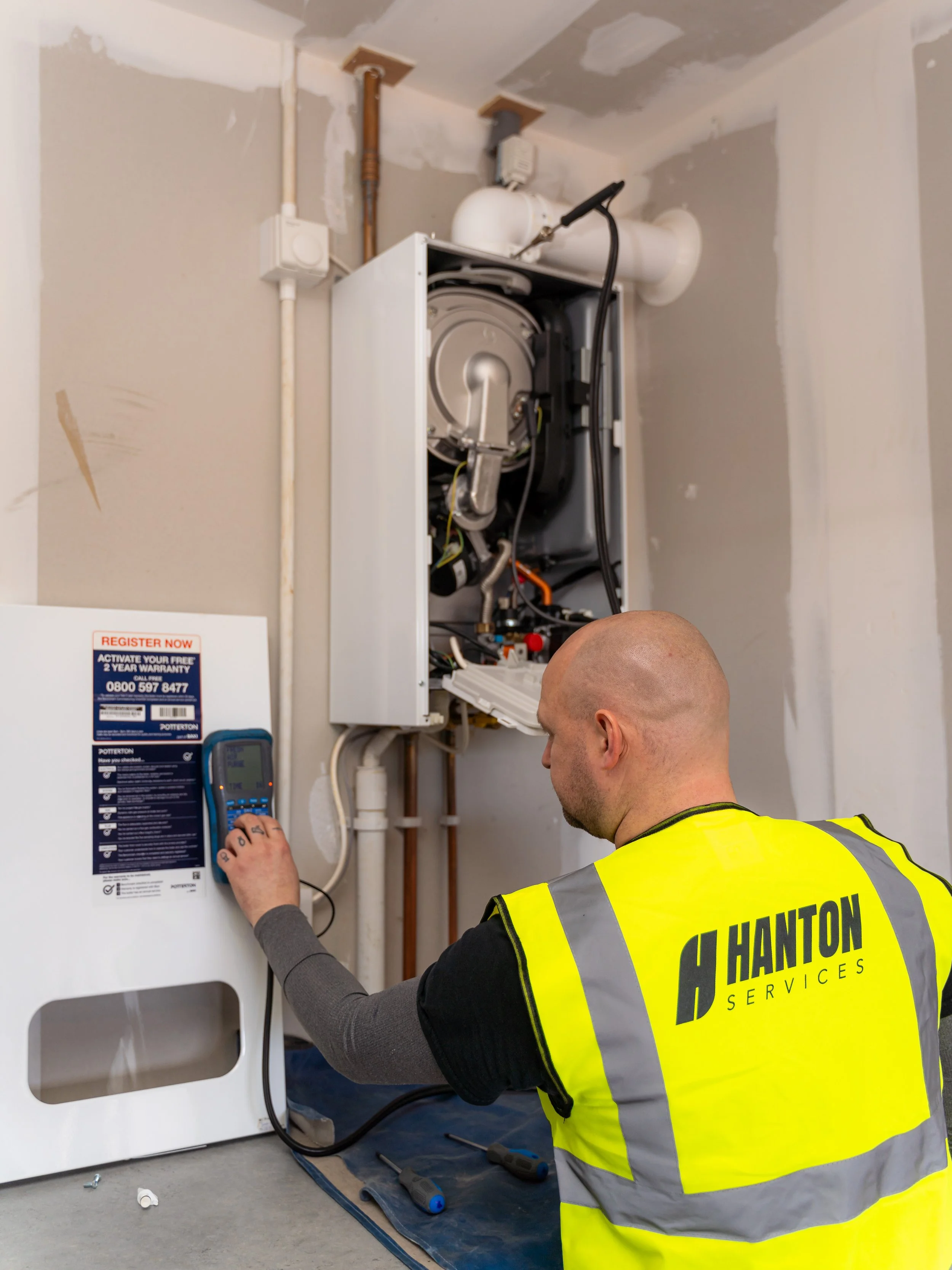 A technician in a yellow safety vest working on an open dishwasher or appliance with internal components, in a room with unfinished walls and visible pipes.