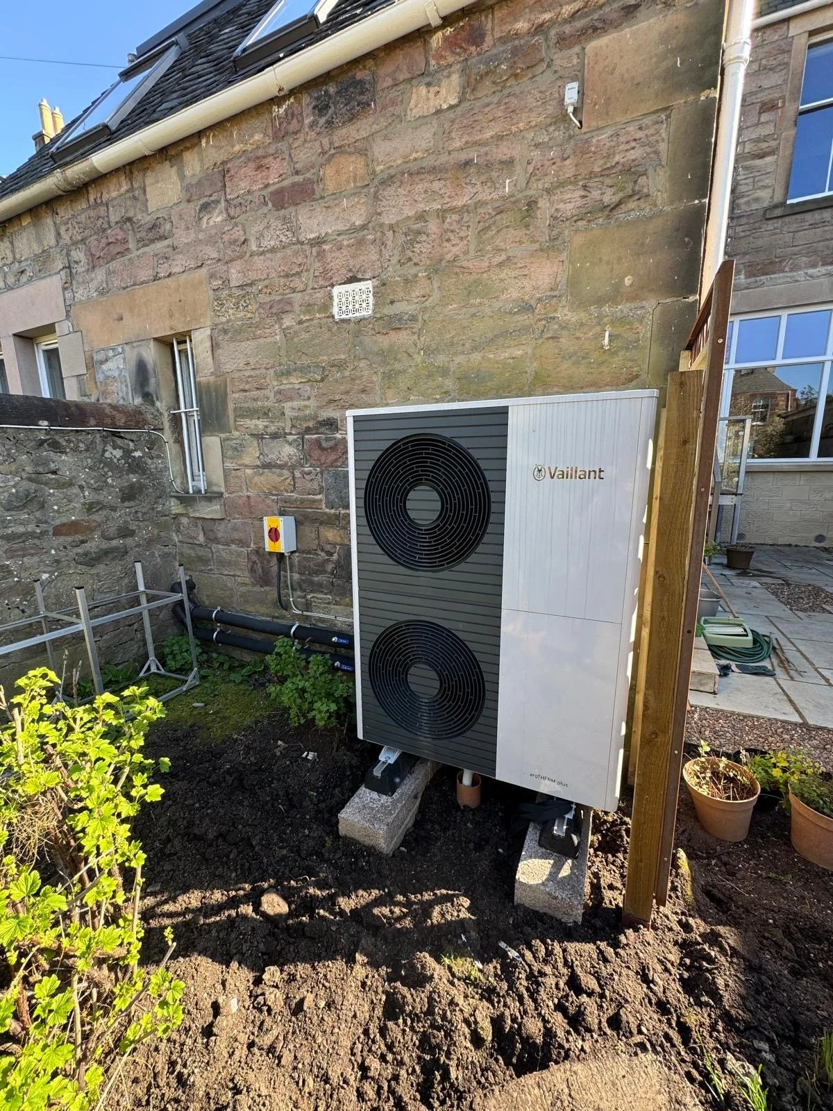 Outdoor air source heat pump with black fans, mounted on cinder blocks in a garden area, next to a stone wall of a building, with plants and garden tools nearby.