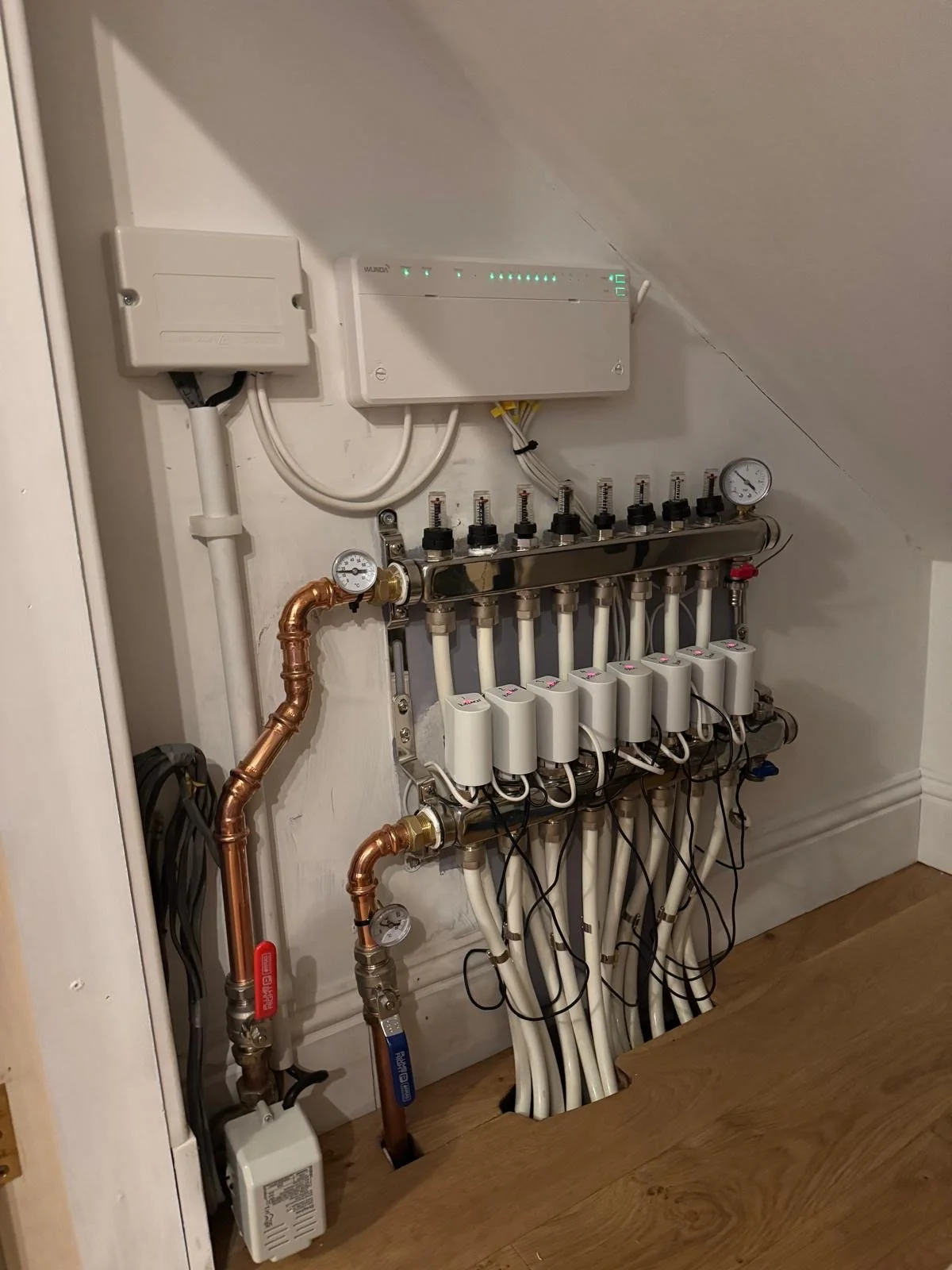 A heating system manifold with multiple pipes, gauges, and electronic controls, mounted on a wall in a room with wooden flooring.