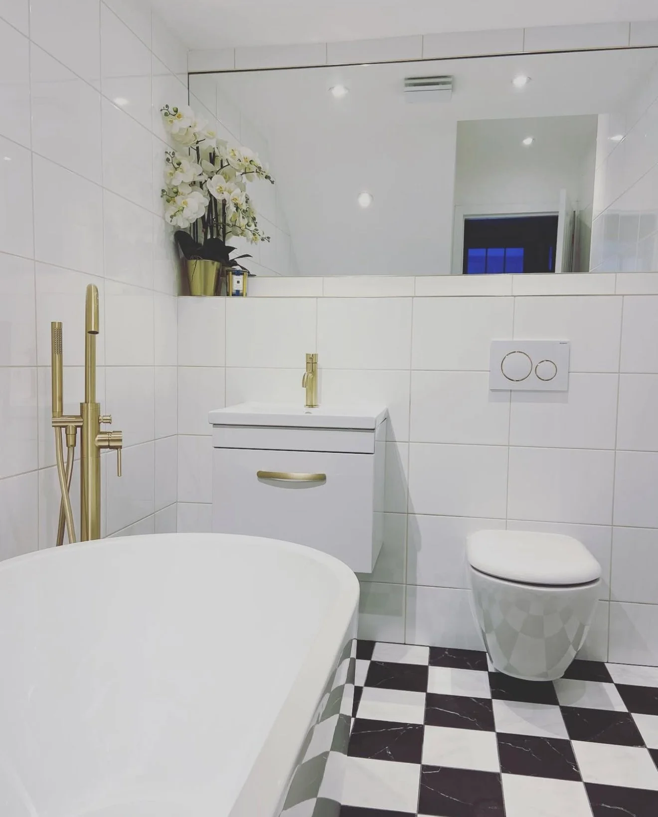 Modern white bathroom with black and white checkered floor, gold fixtures, wall-mounted toilet, small white cabinet, large mirror, and white orchids in a gold pot.