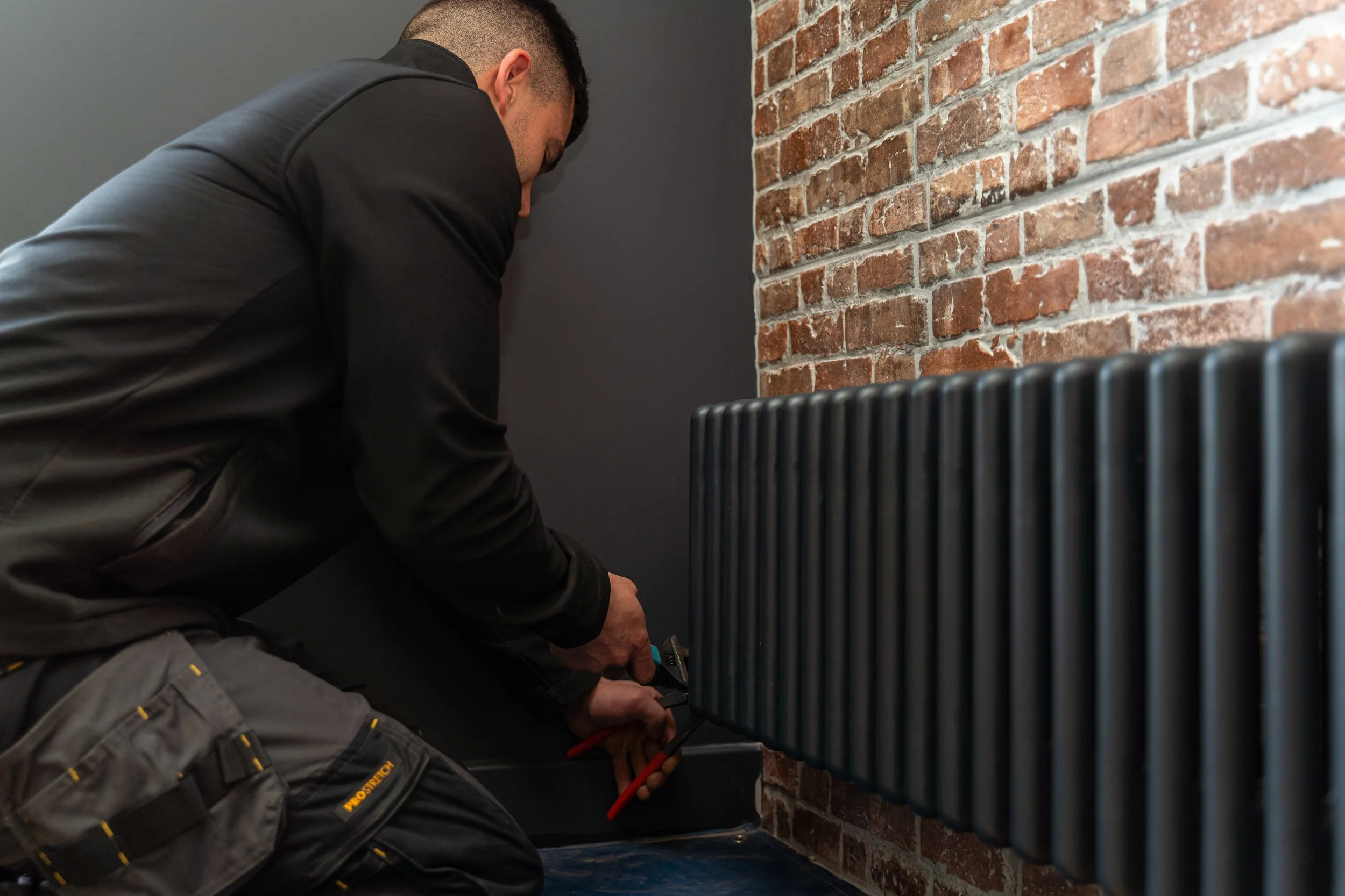 A person wearing a black jacket and gray work pants is kneeling down and working on a black radiator or heater next to a brick wall with a gray wall in the background.