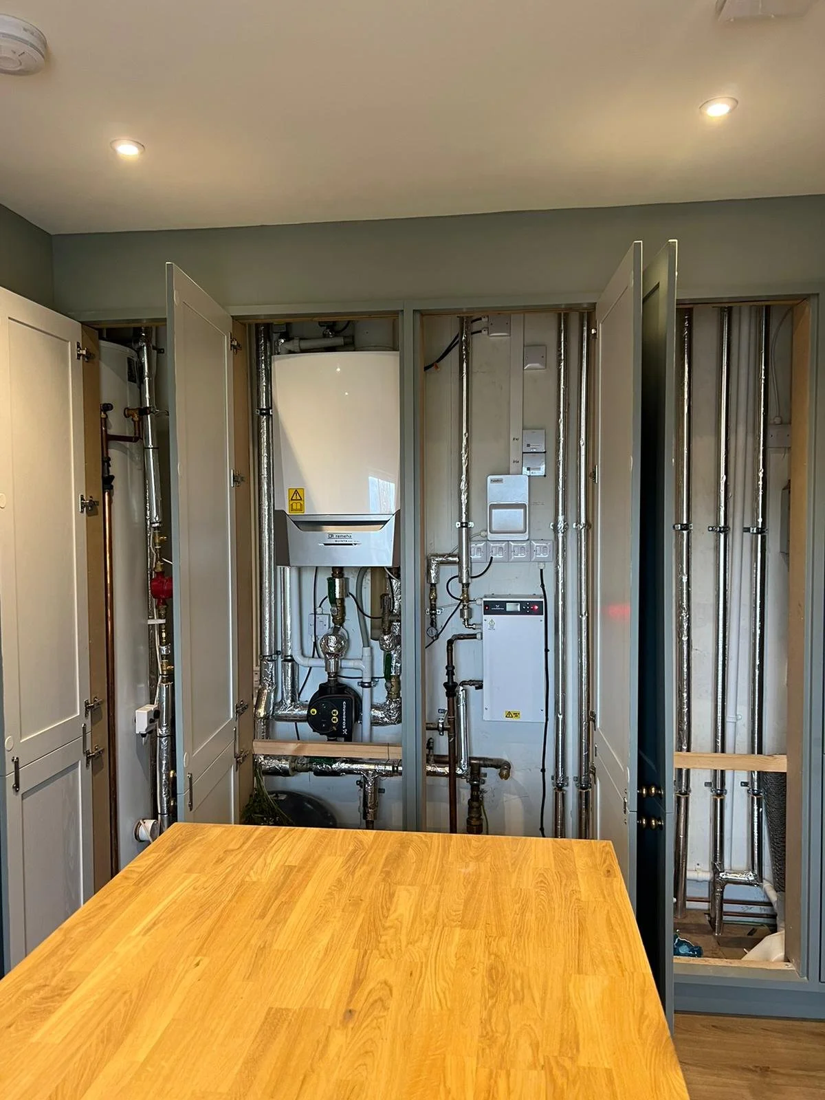 Kitchen with exposed plumbing and electrical wiring behind open cabinet doors with a wooden table in the foreground.