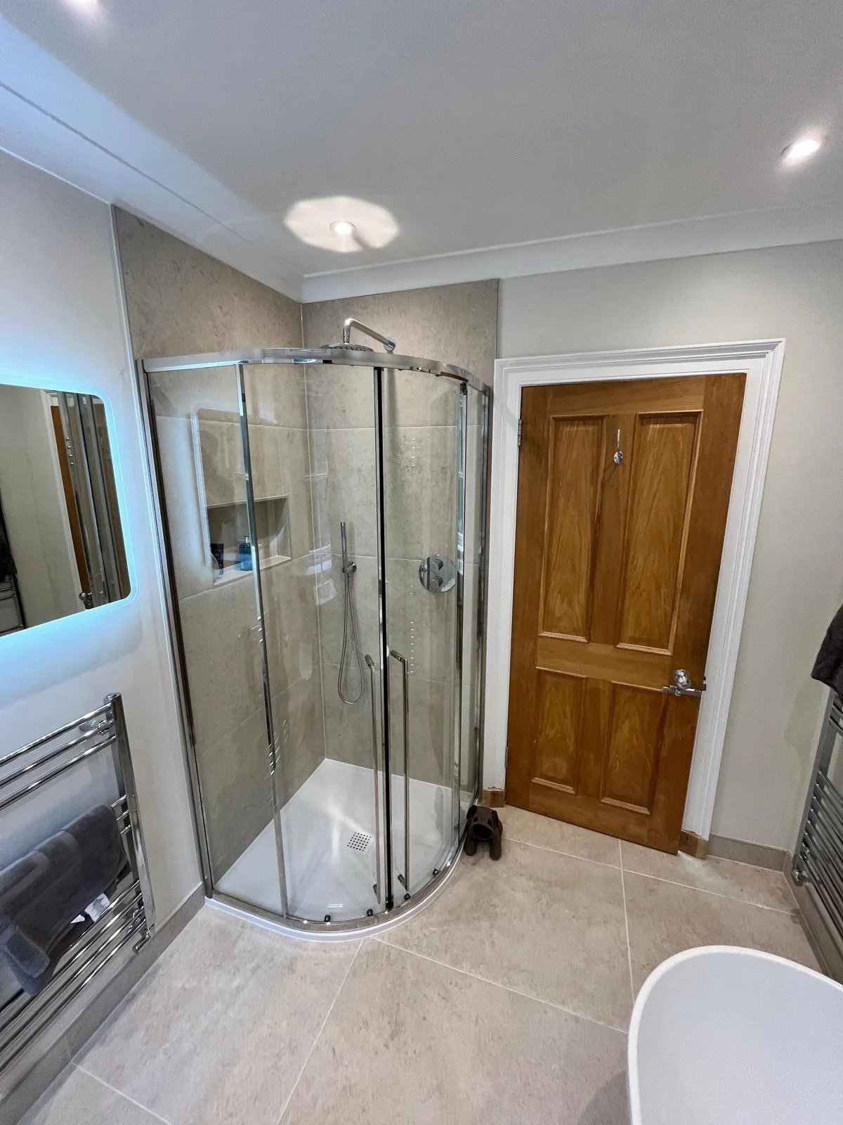 A clean bathroom with a glass shower enclosure, a wooden door, a towel rack with towels, a mirror with a blue backlight, and a partial view of a white bathtub.