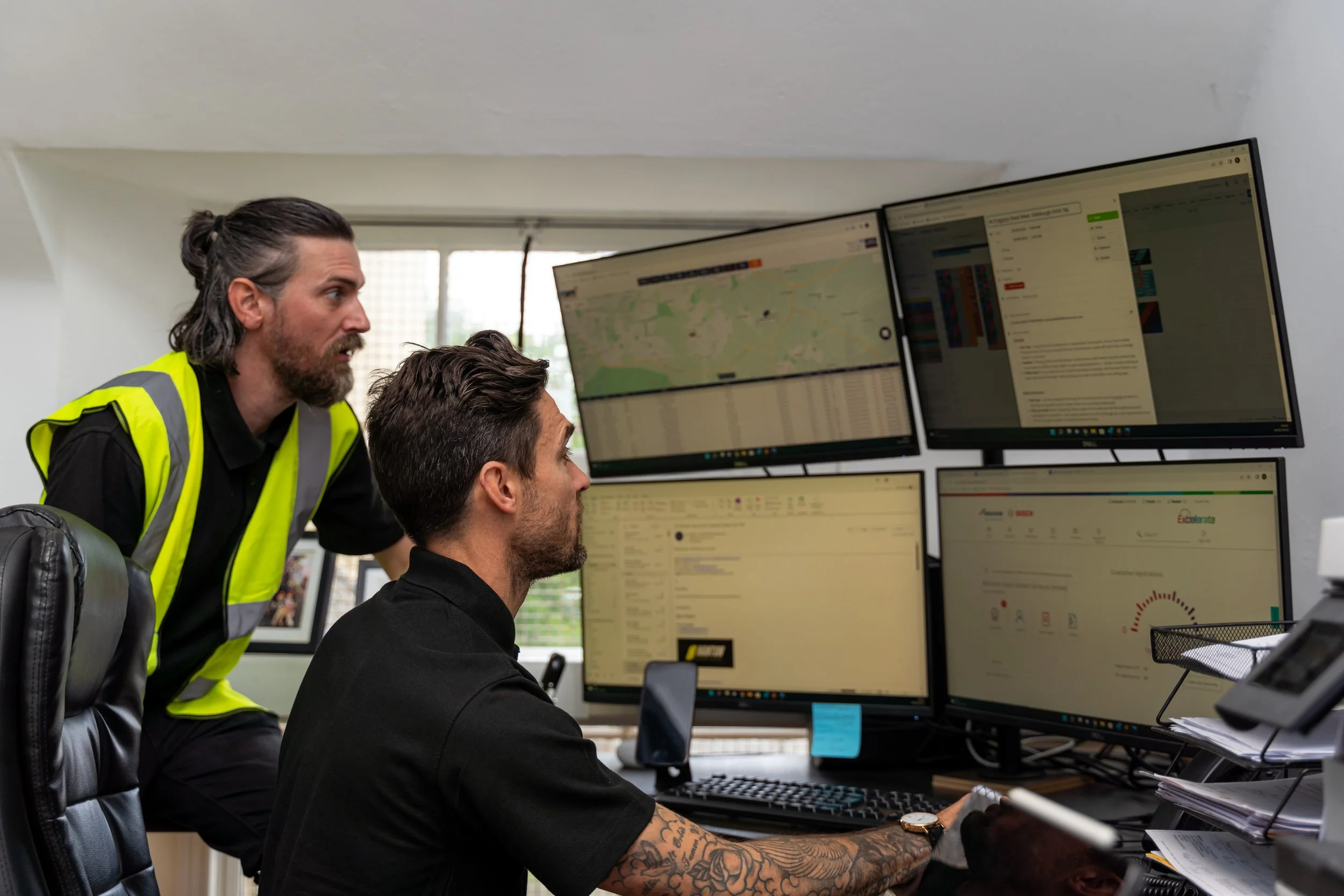 Two men working at a desk with four large computer monitors displaying maps and data, one standing and one seated, in an office with a window.