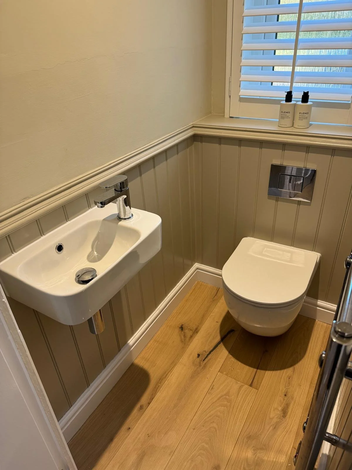 A small bathroom with a wall-mounted toilet, a tiny white sink with a chrome faucet, and a window with white shutters. Two bottles of Elemis skincare products are on the window sill, and the walls are painted beige with beadboard paneling halfway up.