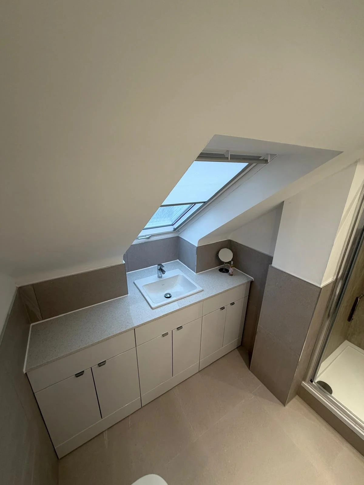 Bathroom with white cabinet, sink, faucet, small mirror, skylight window, tiled walls, and shower.