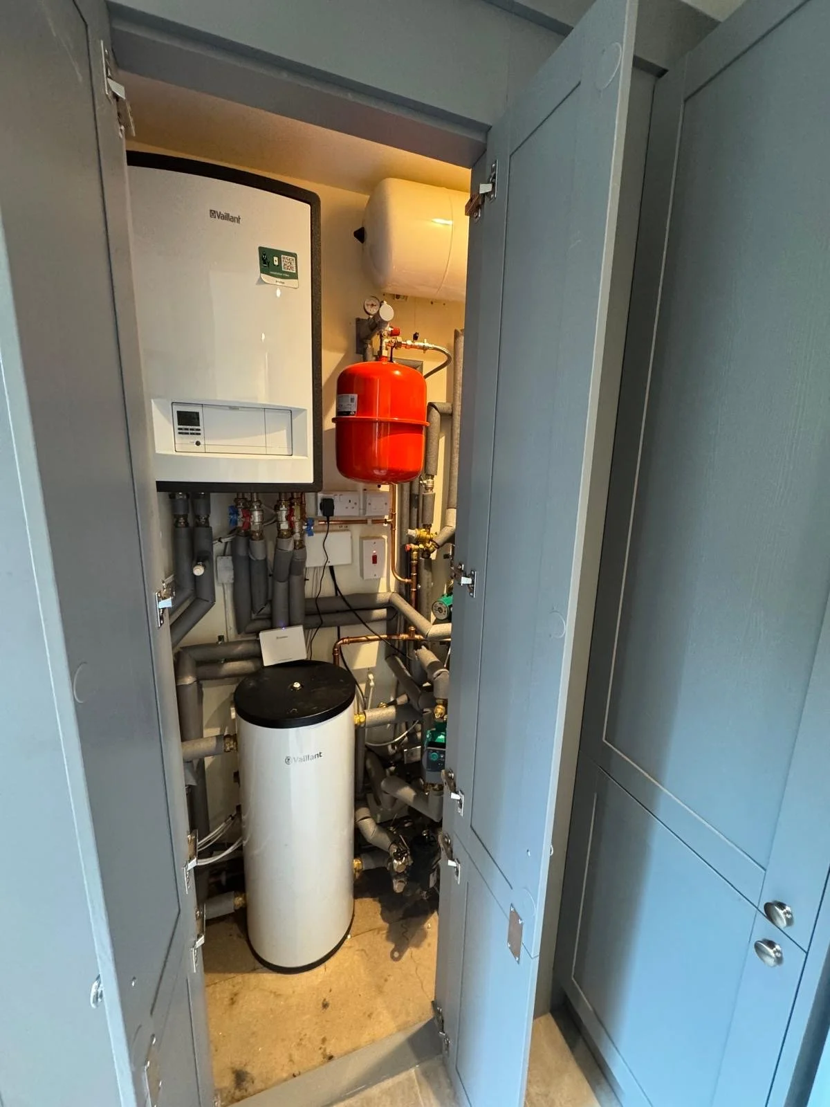 View inside a utility closet showing a boiler, a red expansion tank, pipes, wires, and a water filter.