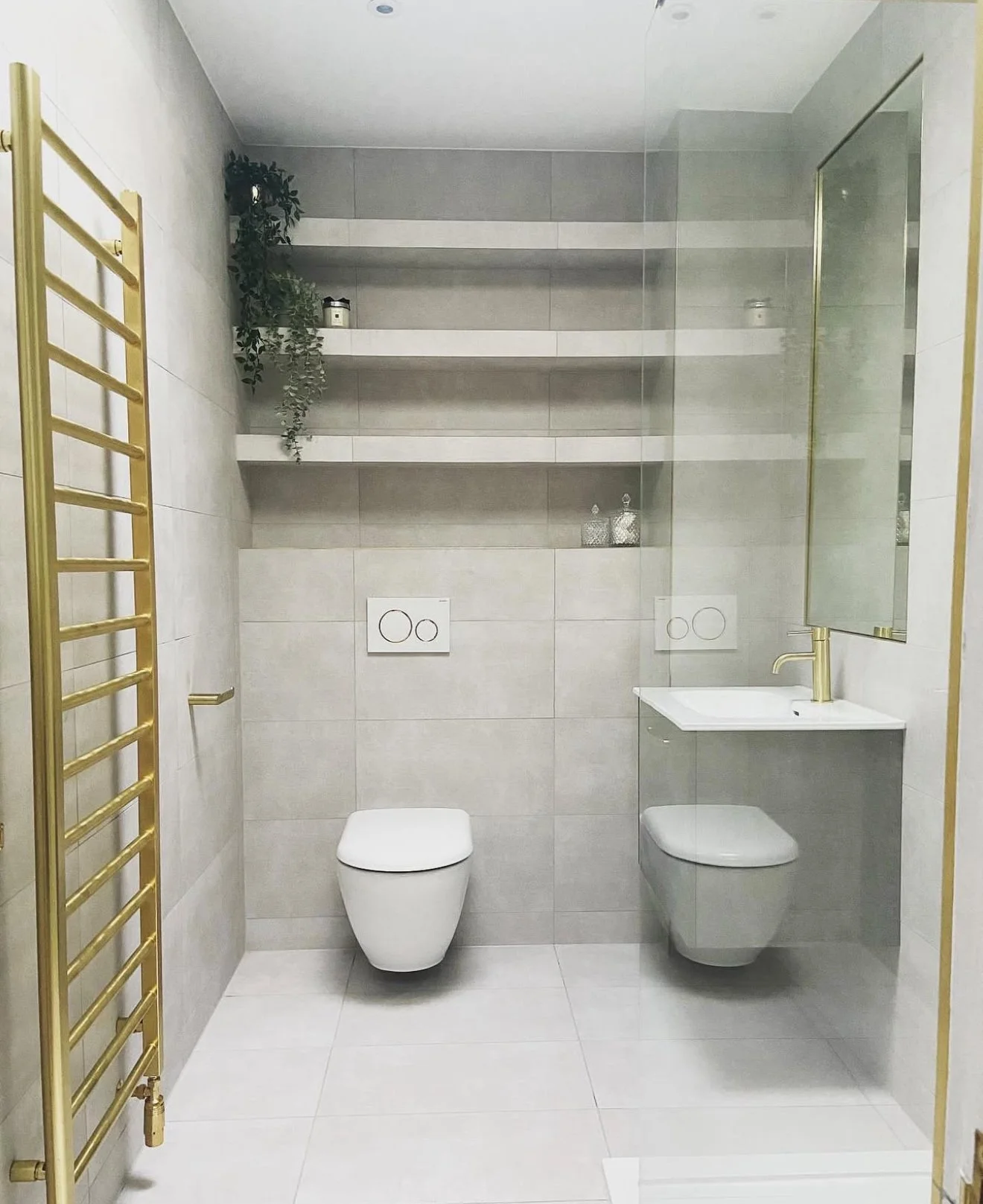 Modern bathroom with two wall-mounted toilets, a mirror, a sink with a gold faucet, glass shower enclosure, gold towel warmer, and open shelves with decorative items and plants.