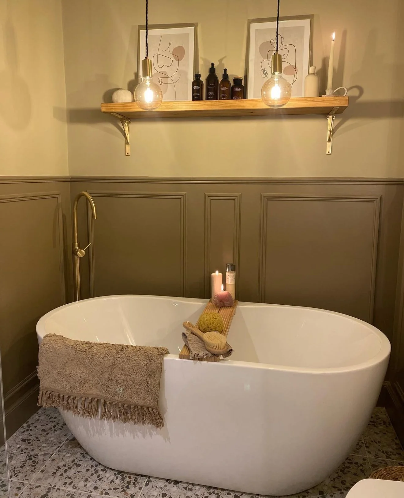 A cozy bathtub scene in a bathroom with beige walls and wainscoting. The white freestanding oval bathtub has a beige towel draped over its edge. On the edge of the tub, there is a wooden tray holding a candle, natural sponge, body brush, and skincare