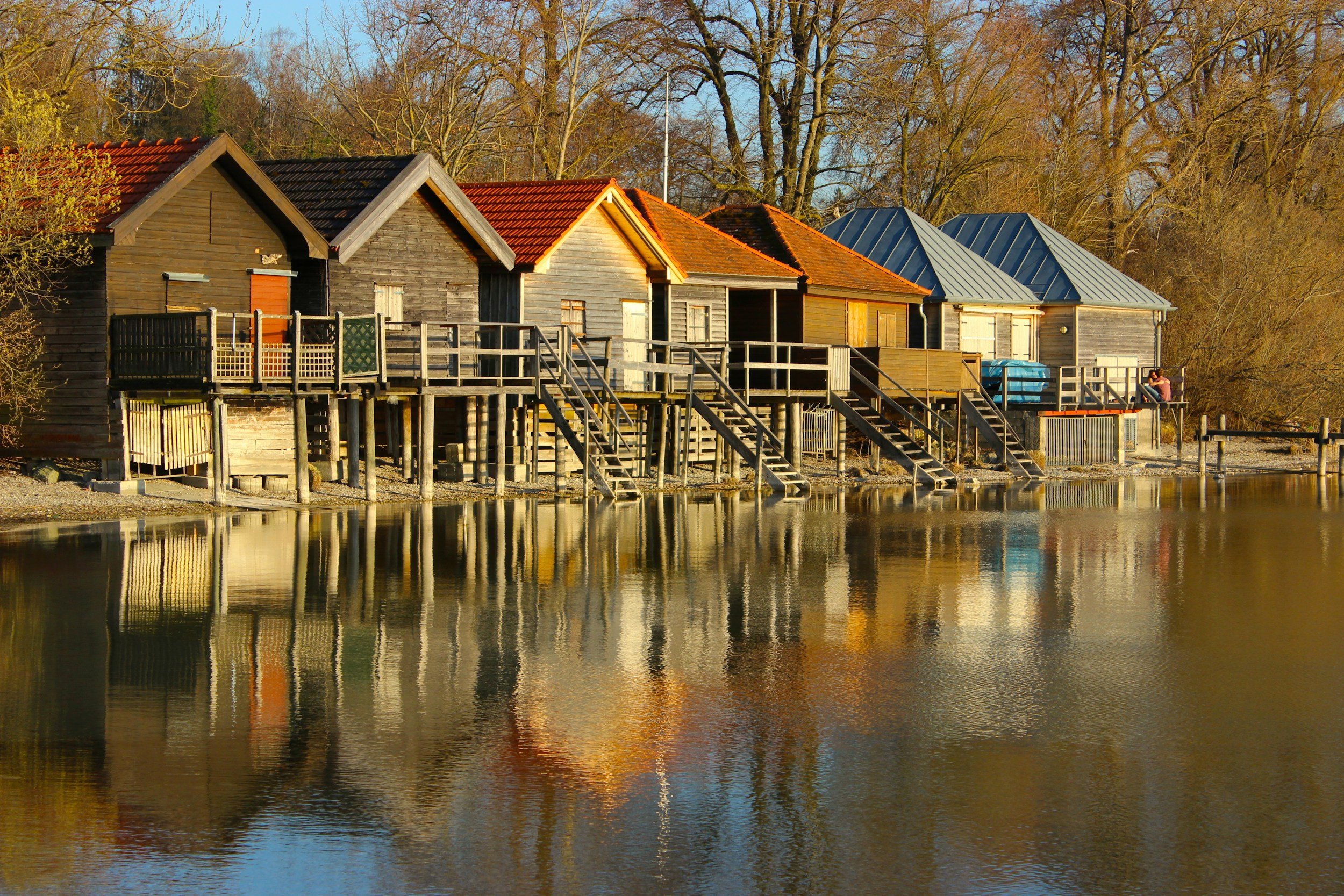 A row of floating wooden houses on a calm body of water, with stairs leading down to the shoreline and trees in the background.