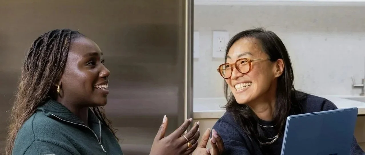 Two women are smiling and chatting in a kitchen. One woman has dark skin, long braids, and is wearing a dark zip-up top. The other woman has light skin, glasses, and shoulder-length black hair, with a laptop on the table in front of her.