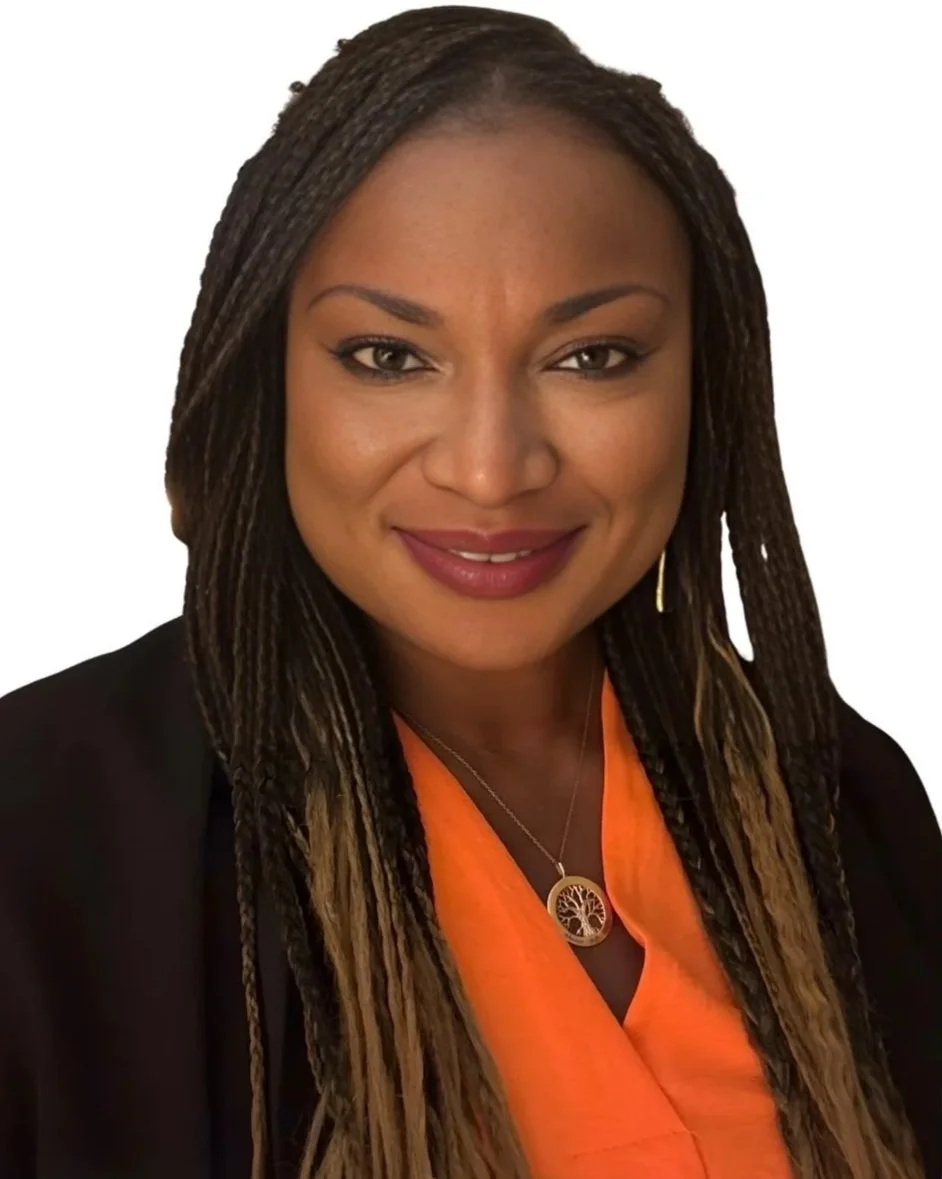 A woman with long braided hair, wearing a black blazer over an orange top, smiling, with a necklace featuring a tree of life pendant, against a plain white background.