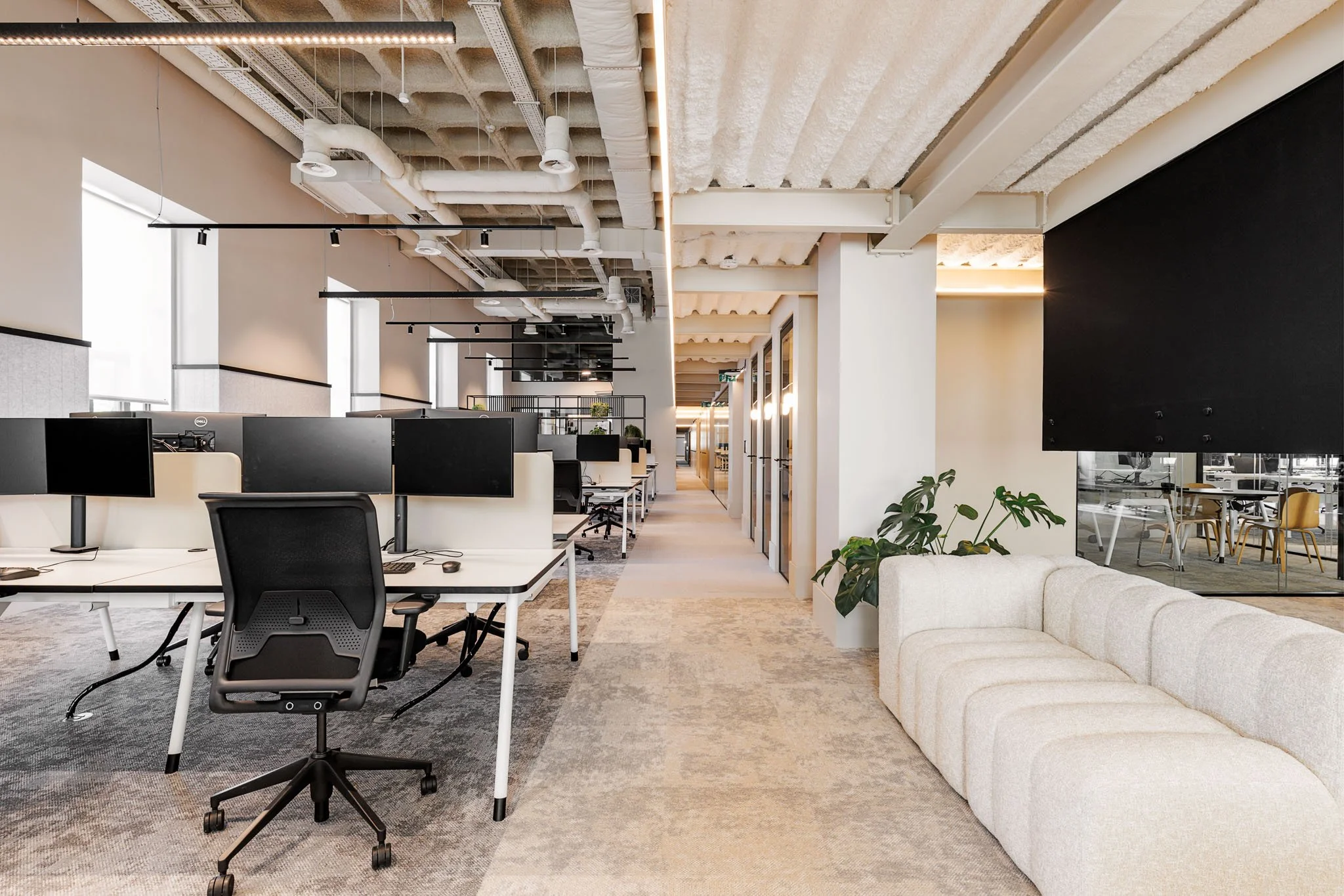 Escritório moderno com estações de trabalho com computadores, cadeira ergonômica, sofá branco, planta e espaço de descanso, com iluminação natural e detalhes em preto e branco.