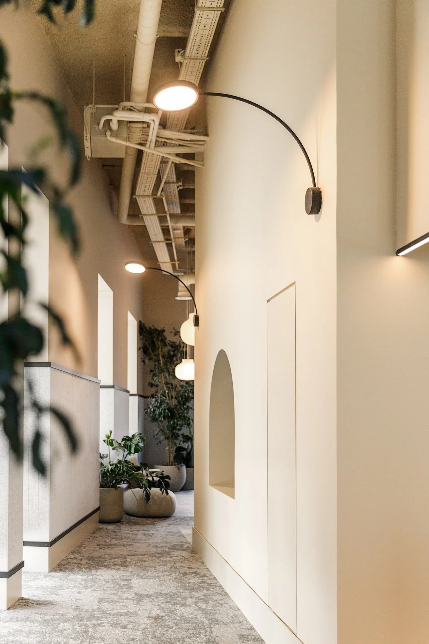 Corredor interno com iluminação moderna, plantas em vasos decorativos, parede clara com janelas e detalhes arquitetônicos.