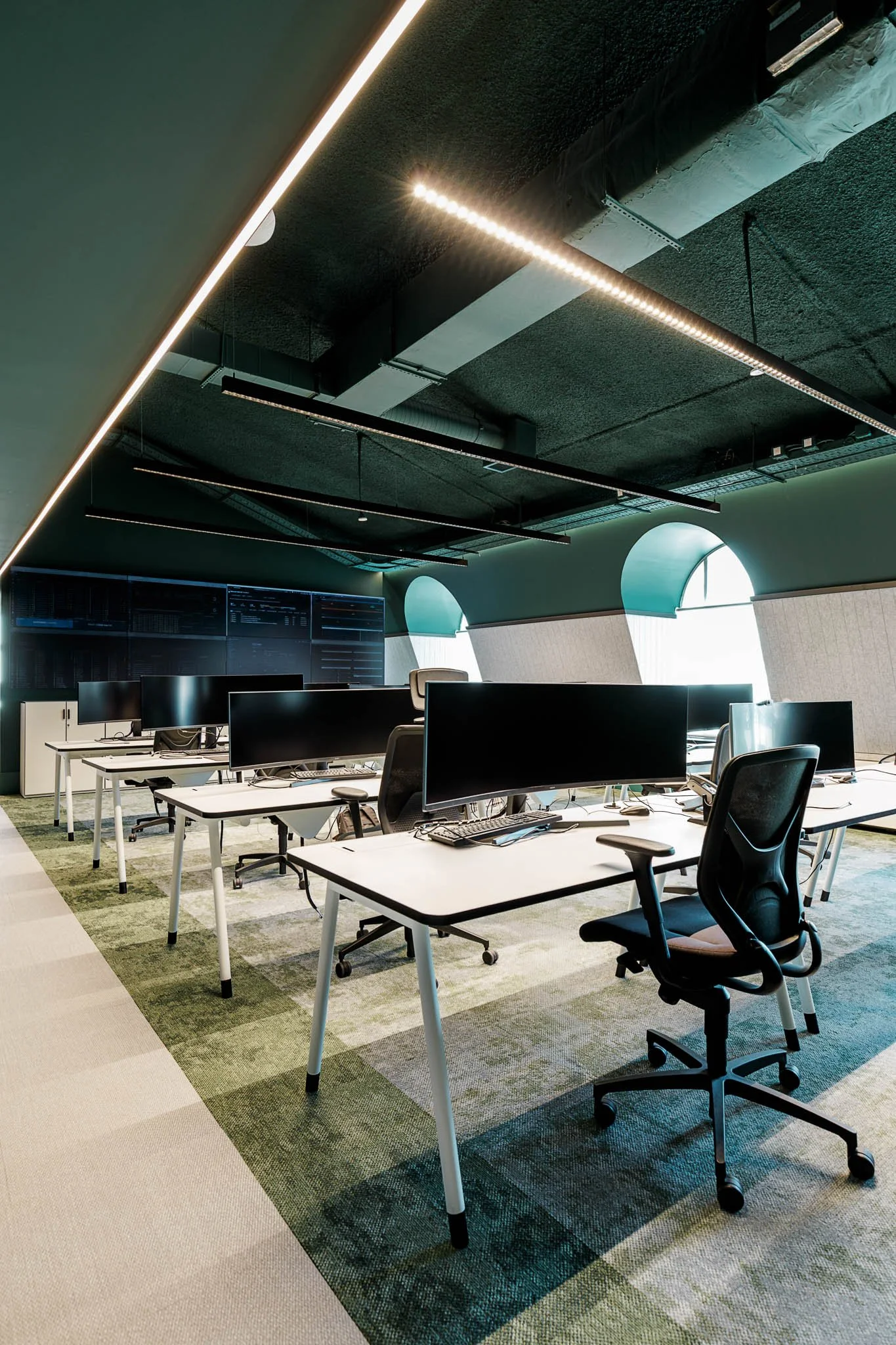 Escritório moderno com várias mesas de trabalho, cadeiras ergonômicas e monitores de computador, iluminação moderna, janelas arqueadas com persianas, piso de carpete com padrão geométrico.