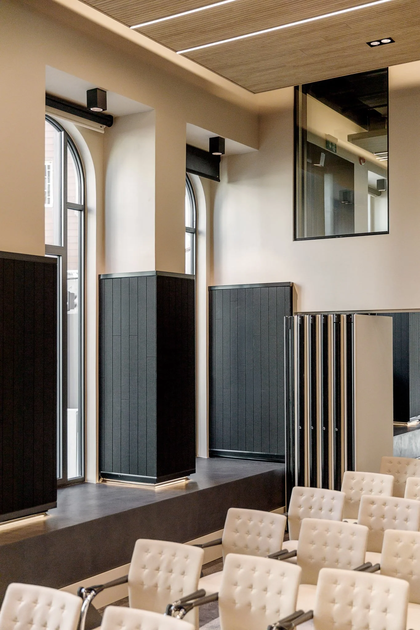 Interior de uma sala com cadeiras brancas e painéis pretos na parede, janelas altas de vidro e uma janela de vidro grande na parede.