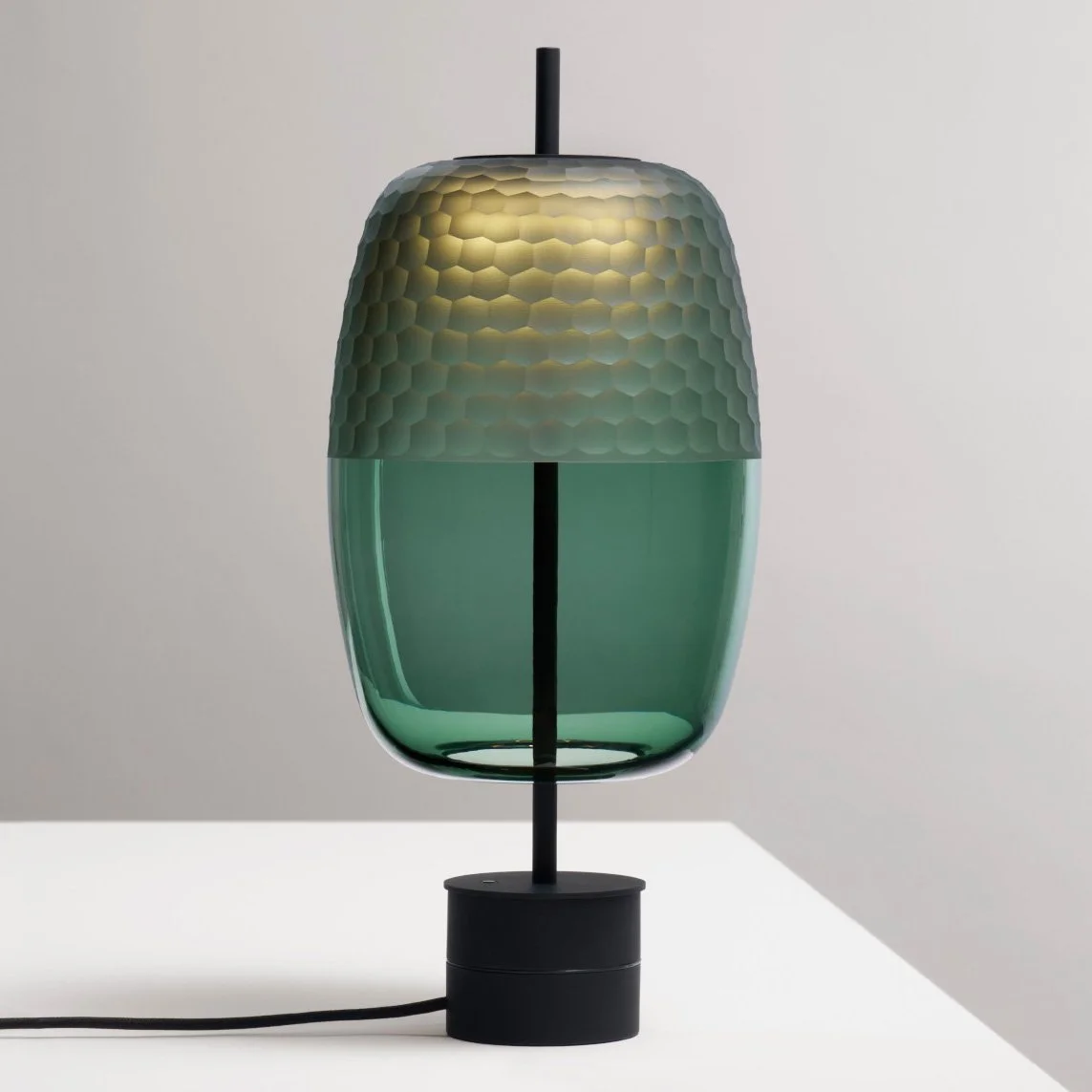 Luminária moderna com design inspirado em um favo de mel, com parte superior em tons dourados e parte inferior em verde translúcido, apoiada por uma base preta e ligada a um fio elétrico.